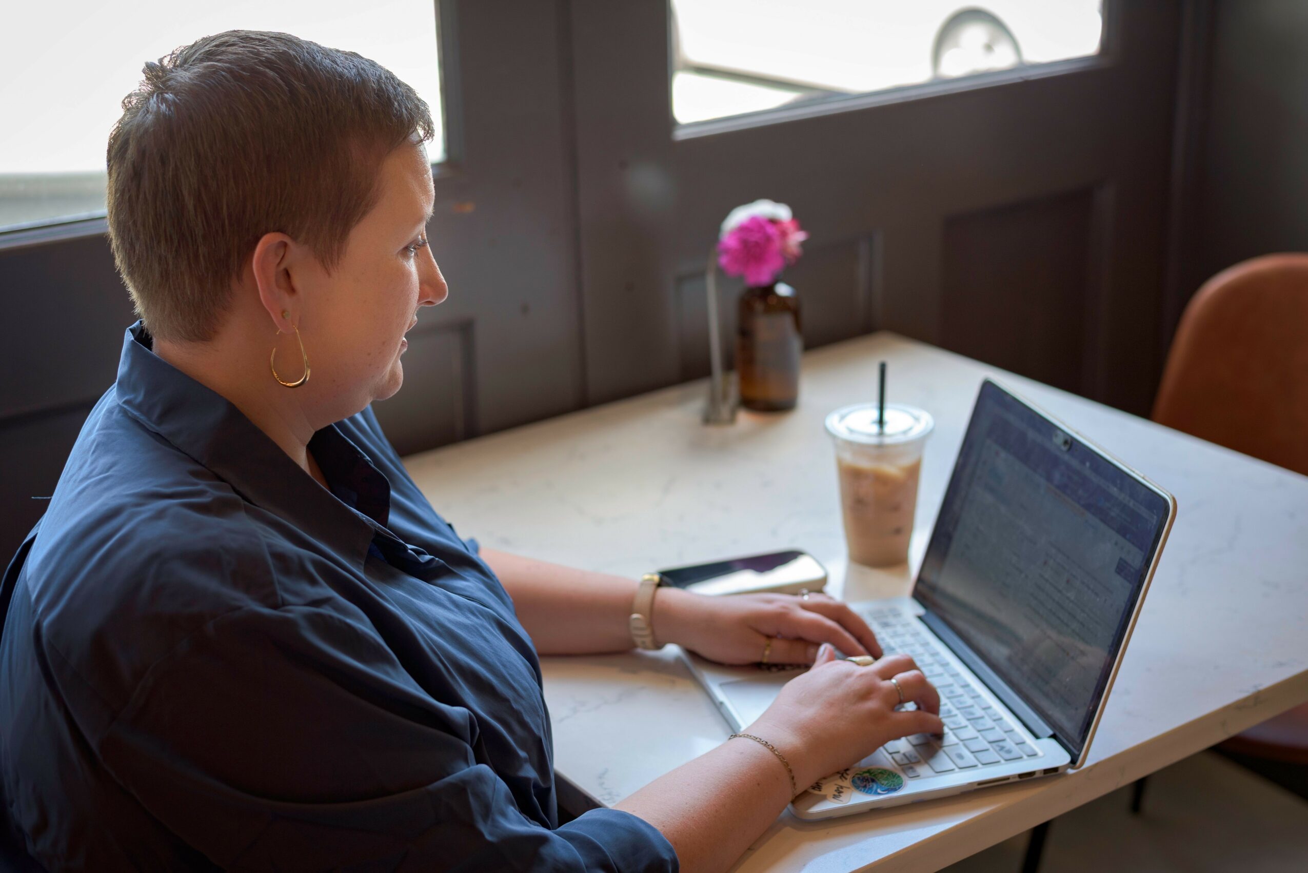 Woman sitting at a table using a laptop, with a drink and a pink flower in a vase nearby.