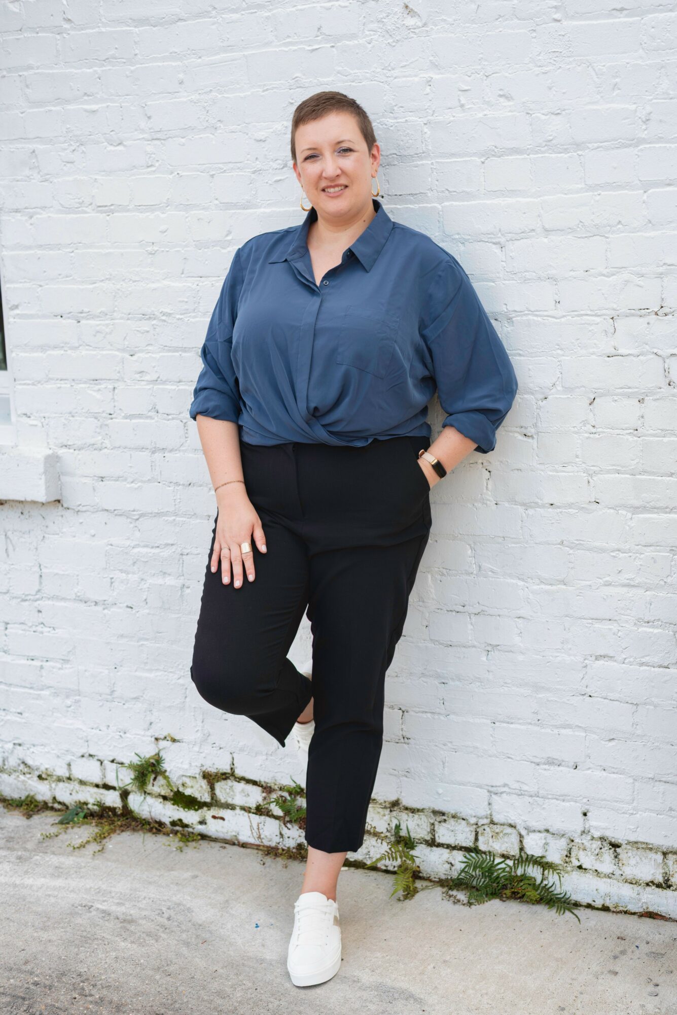 Person standing against a white brick wall, wearing a blue shirt, black pants, and white shoes, smiling.