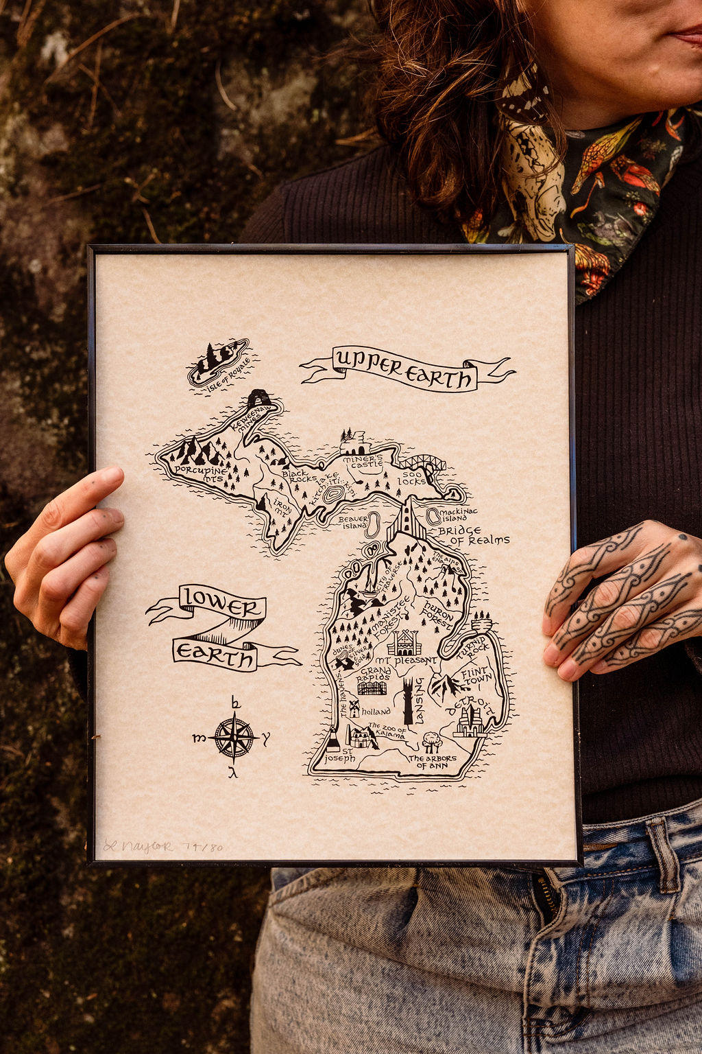Person holding a framed map of Middle-earth with labeled regions, outdoors with trees in background.