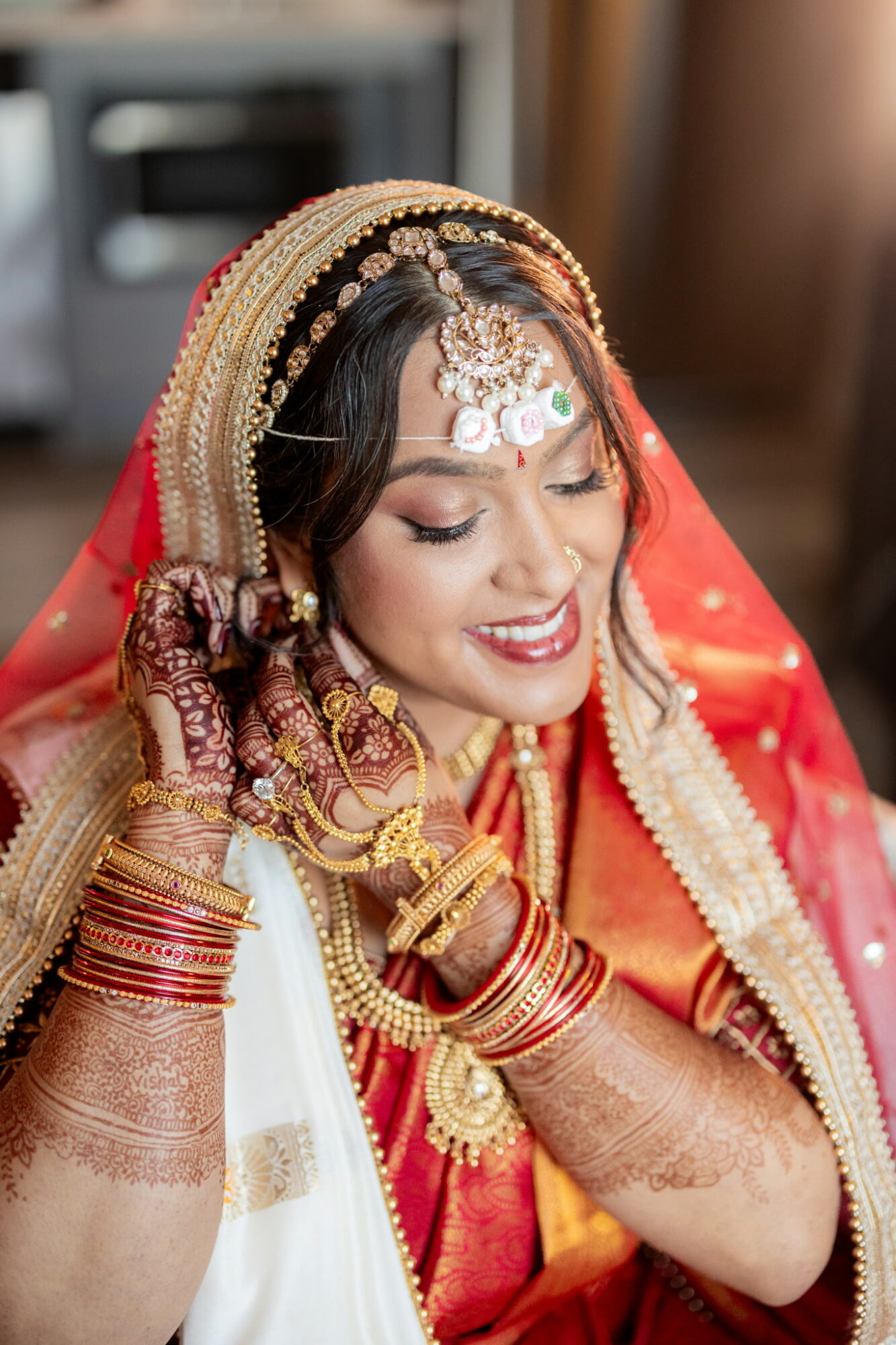 Bride in traditional attire with jewelry, smiling, adjusting her bangles, with a blurred background.
