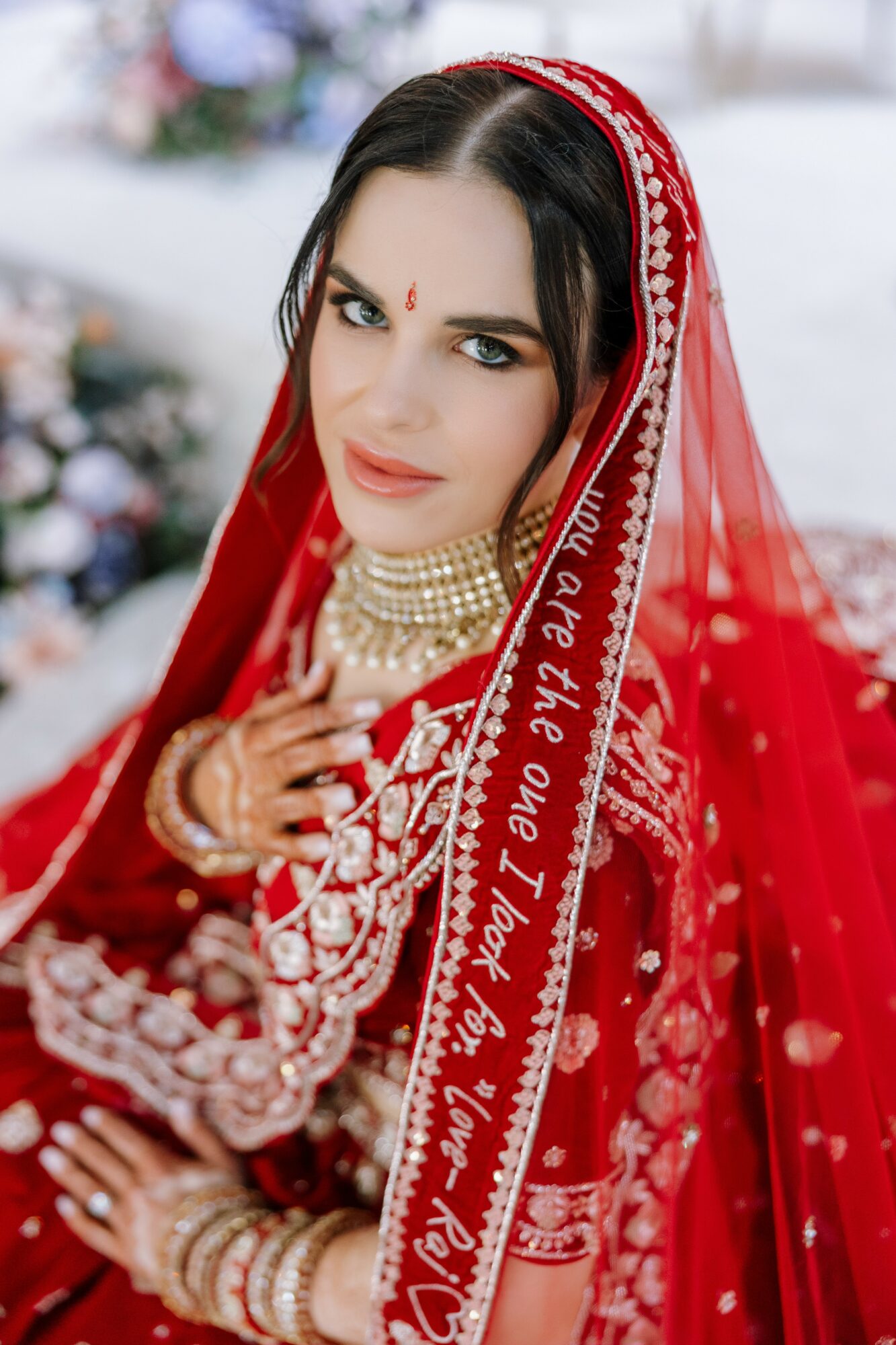 Woman in traditional red attire with jewelry, looking at camera, outdoors with blurred background.
