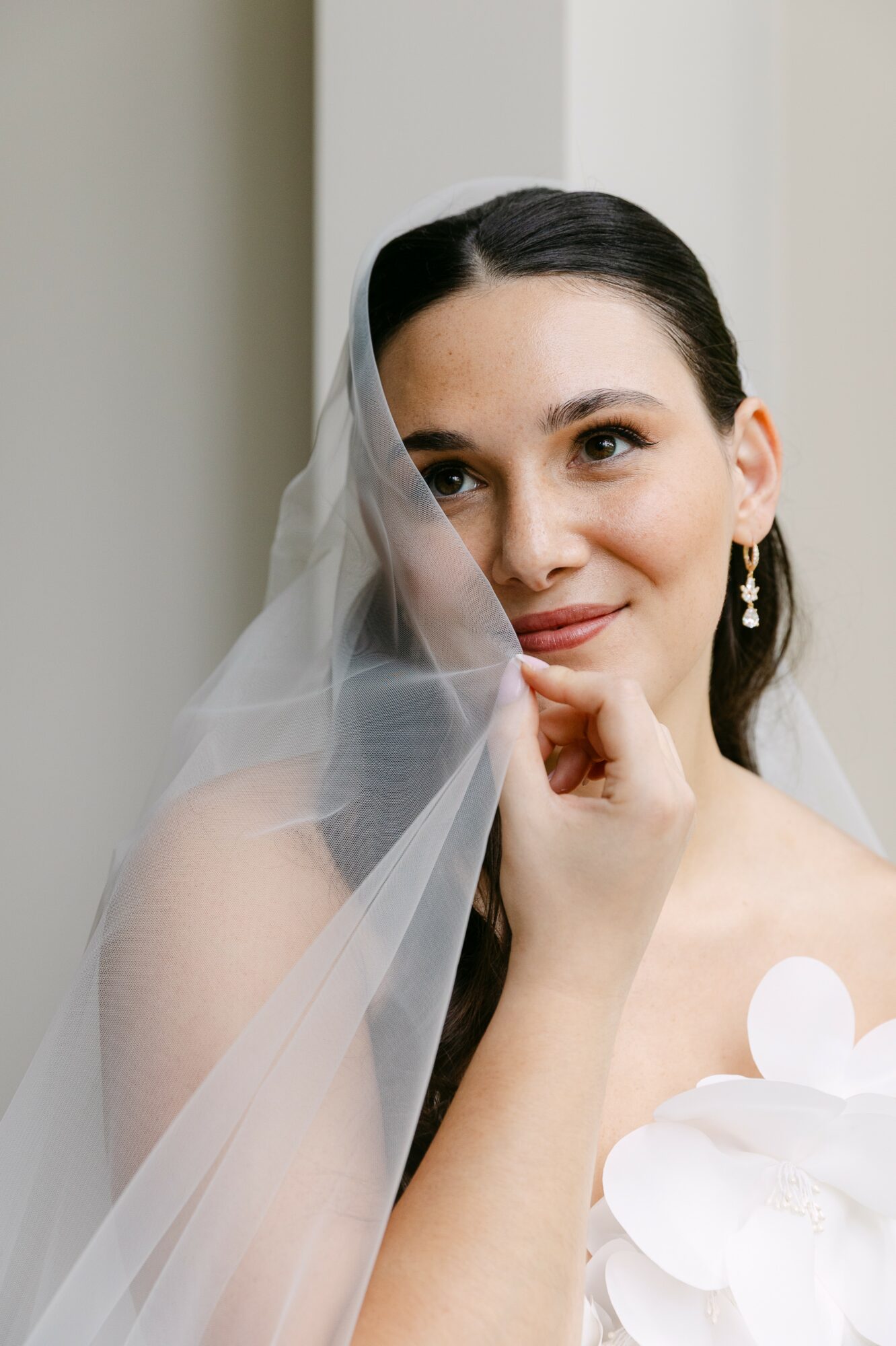 A woman with dark hair and earrings, partially covered by a sheer veil, smiling gently, holding her chin with her hand.