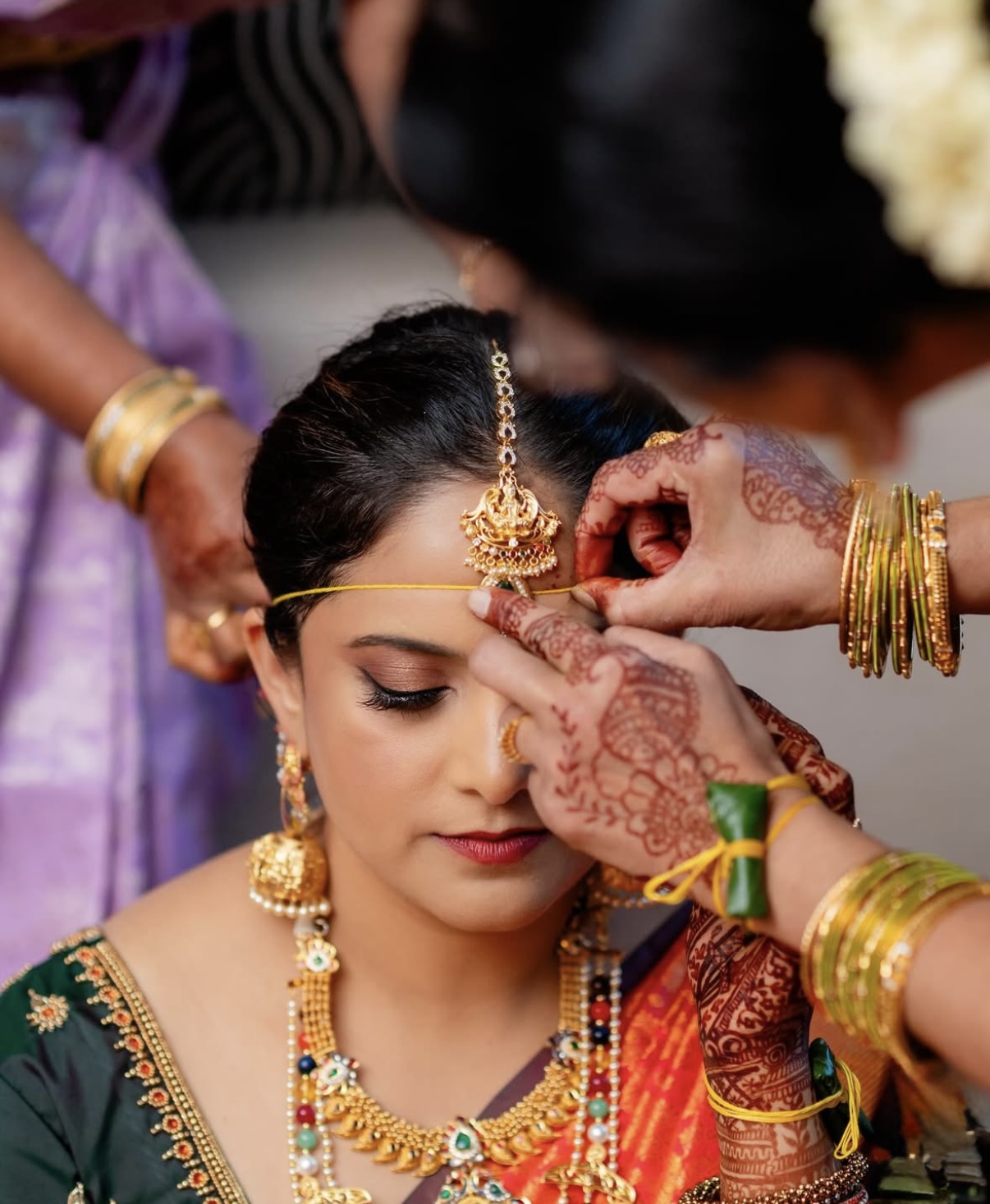 Woman with jewelry having her forehead decorated by a person with hennaed hands, others in traditional attire in background.