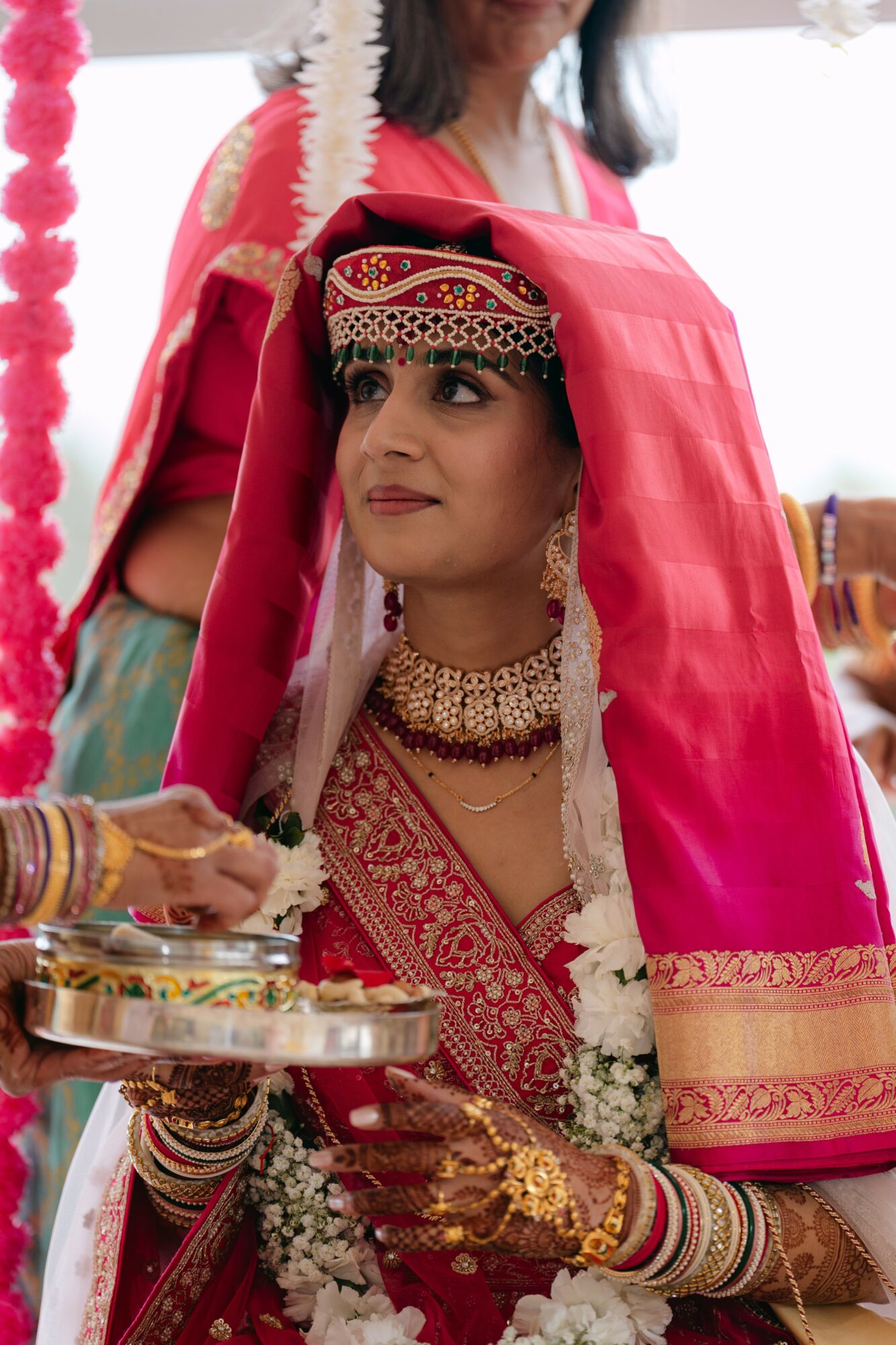 Woman dressed in traditional attire with jewelry, sitting with a pink veil over her head, holding a tray of offerings.
