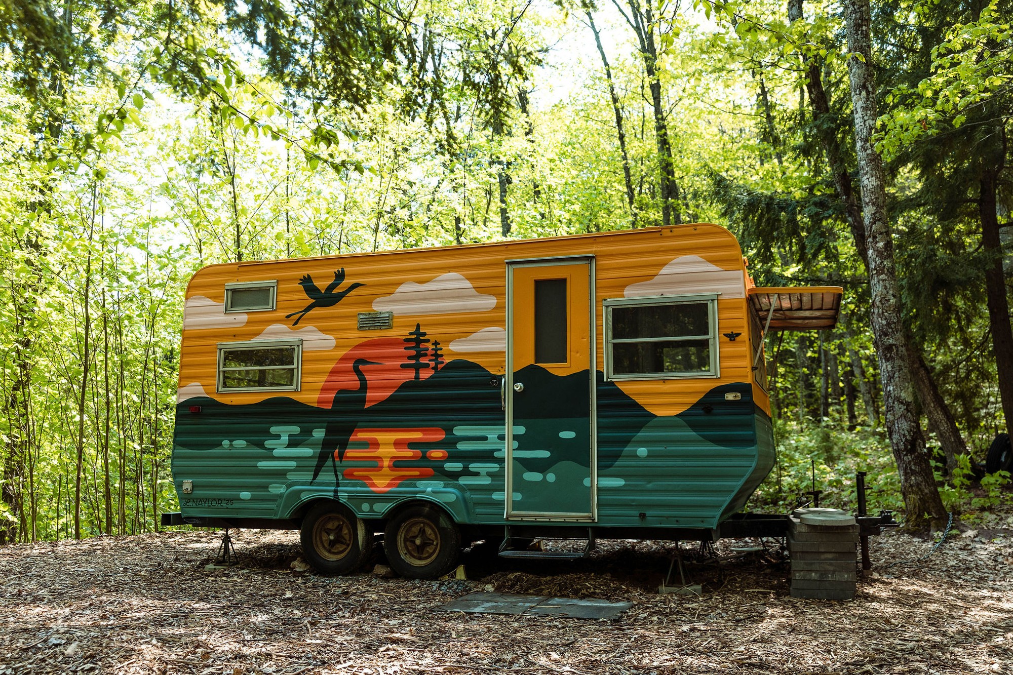 Colorful camper with a mountain and sunset scene, surrounded by trees in a forest, with a small black trash bin nearby.