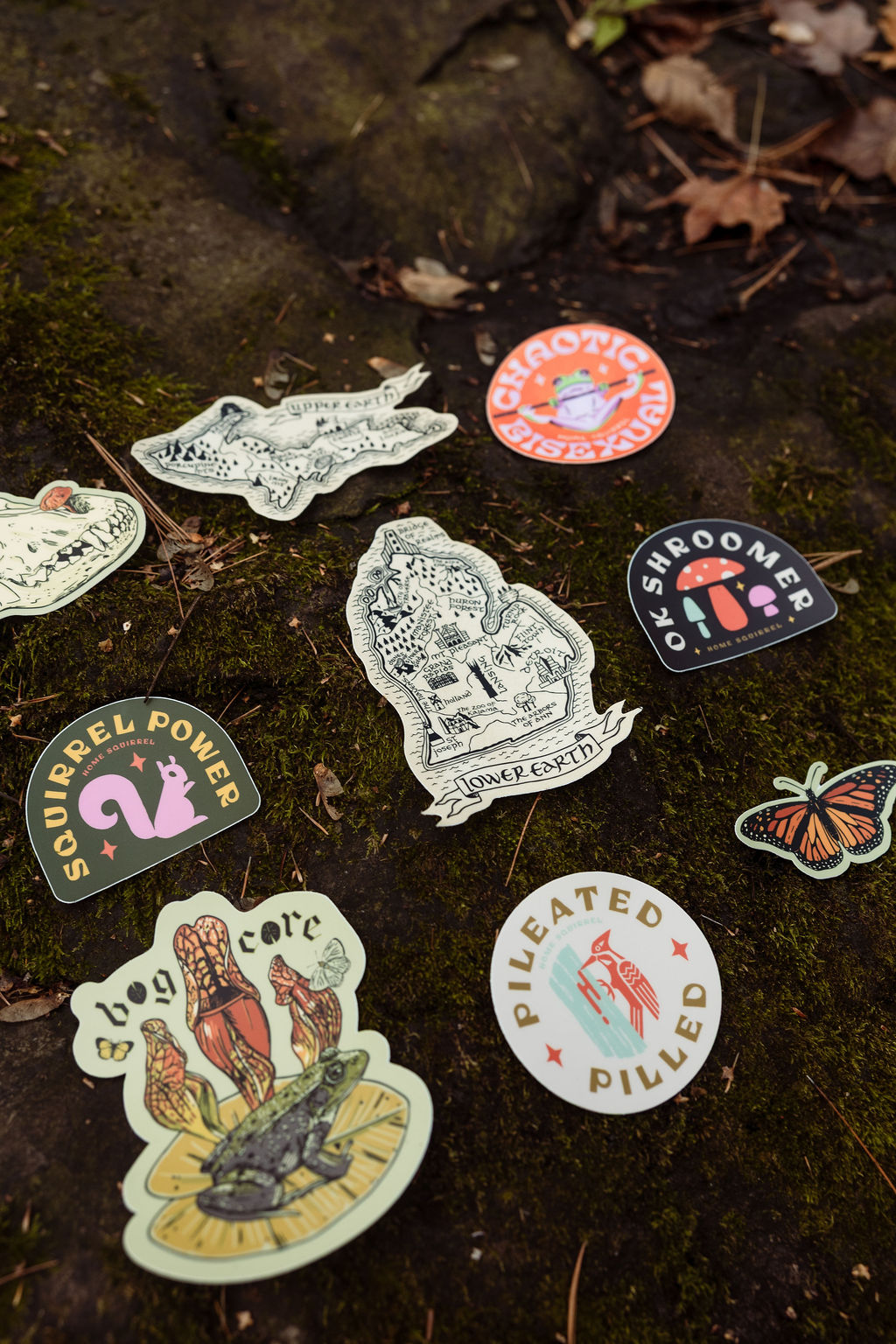 Collection of stickers on mossy ground, including a fish, a butterfly, a mushroom, and circular designs.