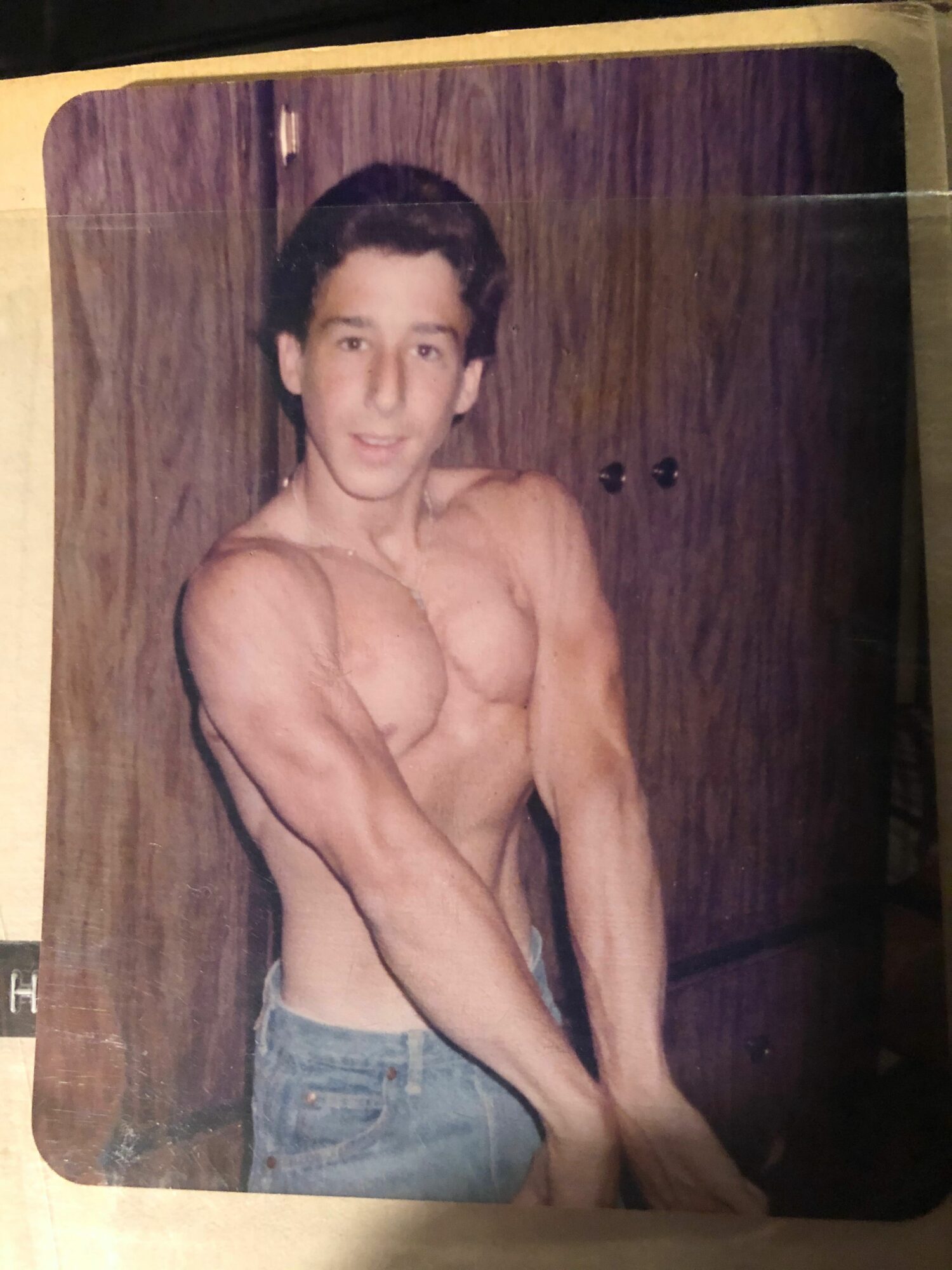 Young man with dark hair, shirtless, sitting on a wooden surface, wearing shorts, looking at the camera.