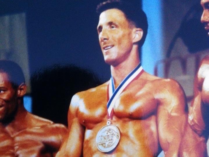 A shirtless man with a medal around his neck smiling on stage, with other shirtless men nearby.