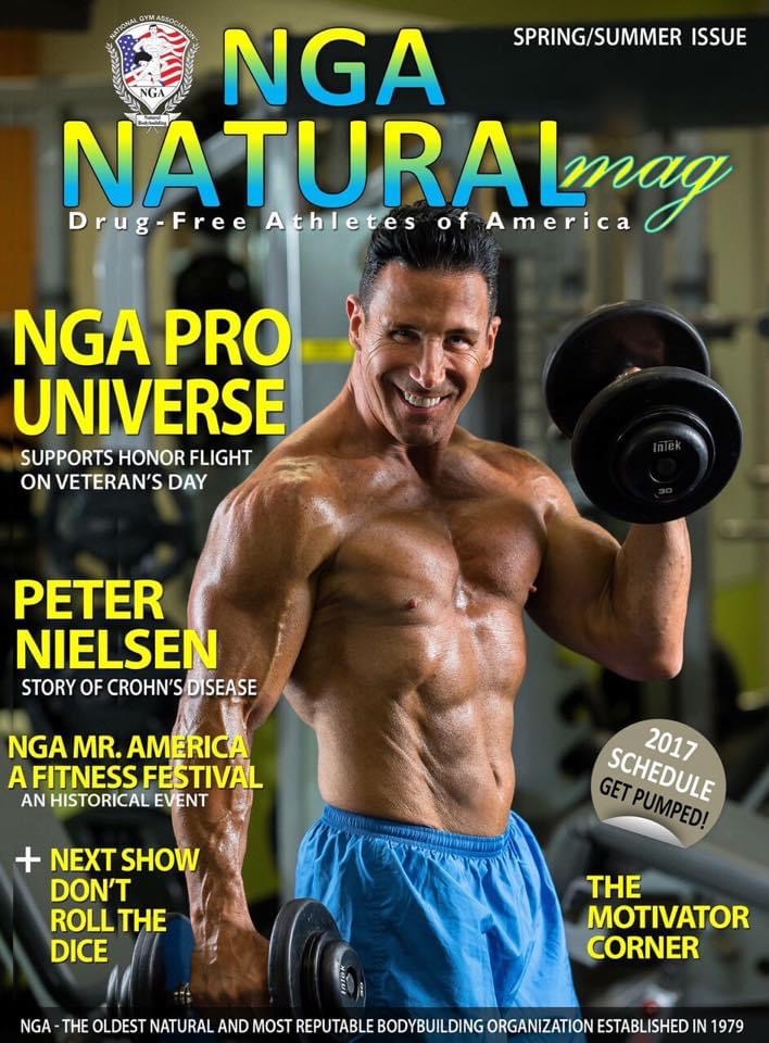 Man lifting dumbbell in gym, smiling, shirtless, wearing blue shorts, background with gym equipment and windows.