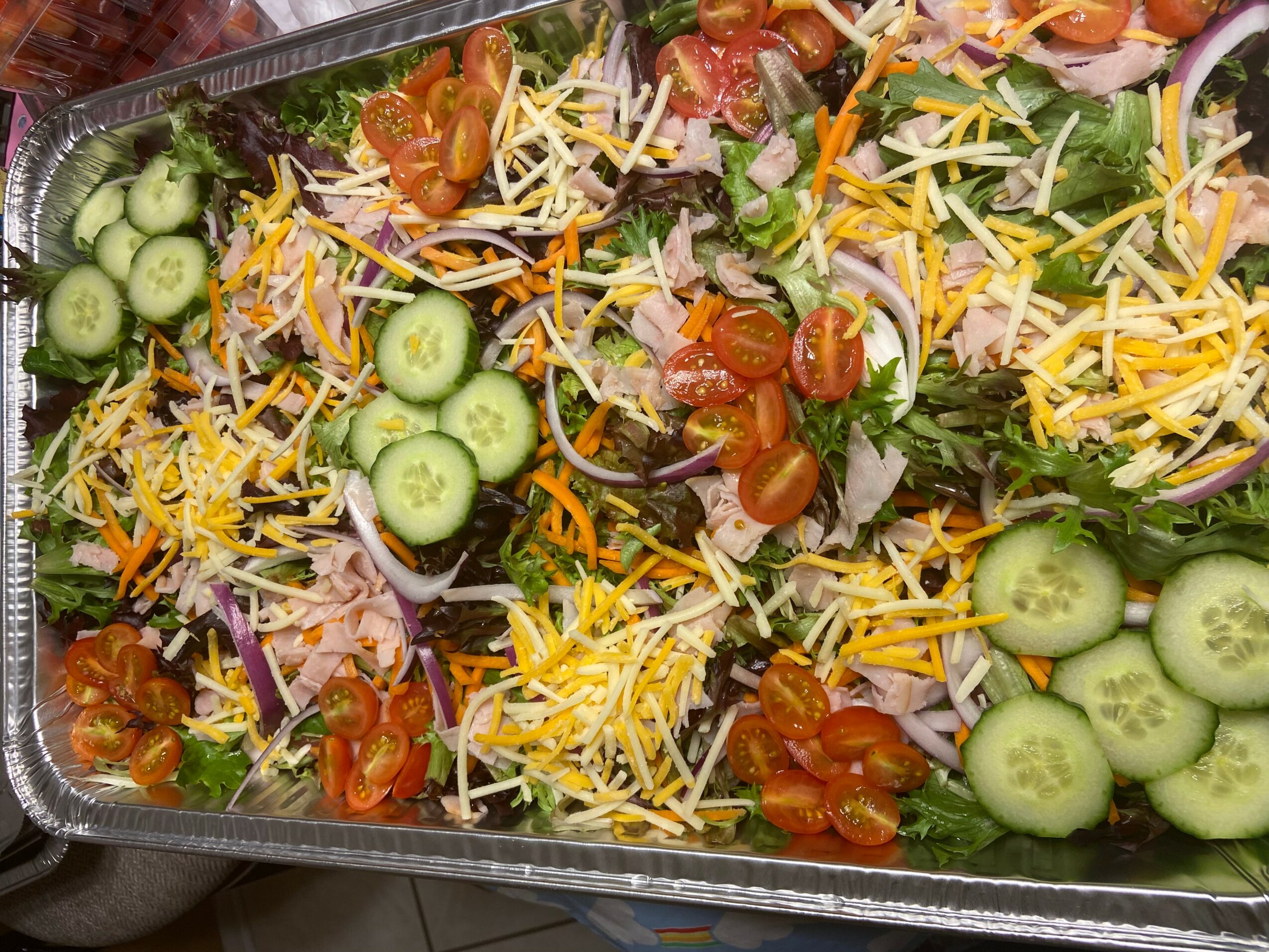 Fresh salad with cucumbers, cherry tomatoes, shredded cheese, lettuce, and shredded carrots in a container.