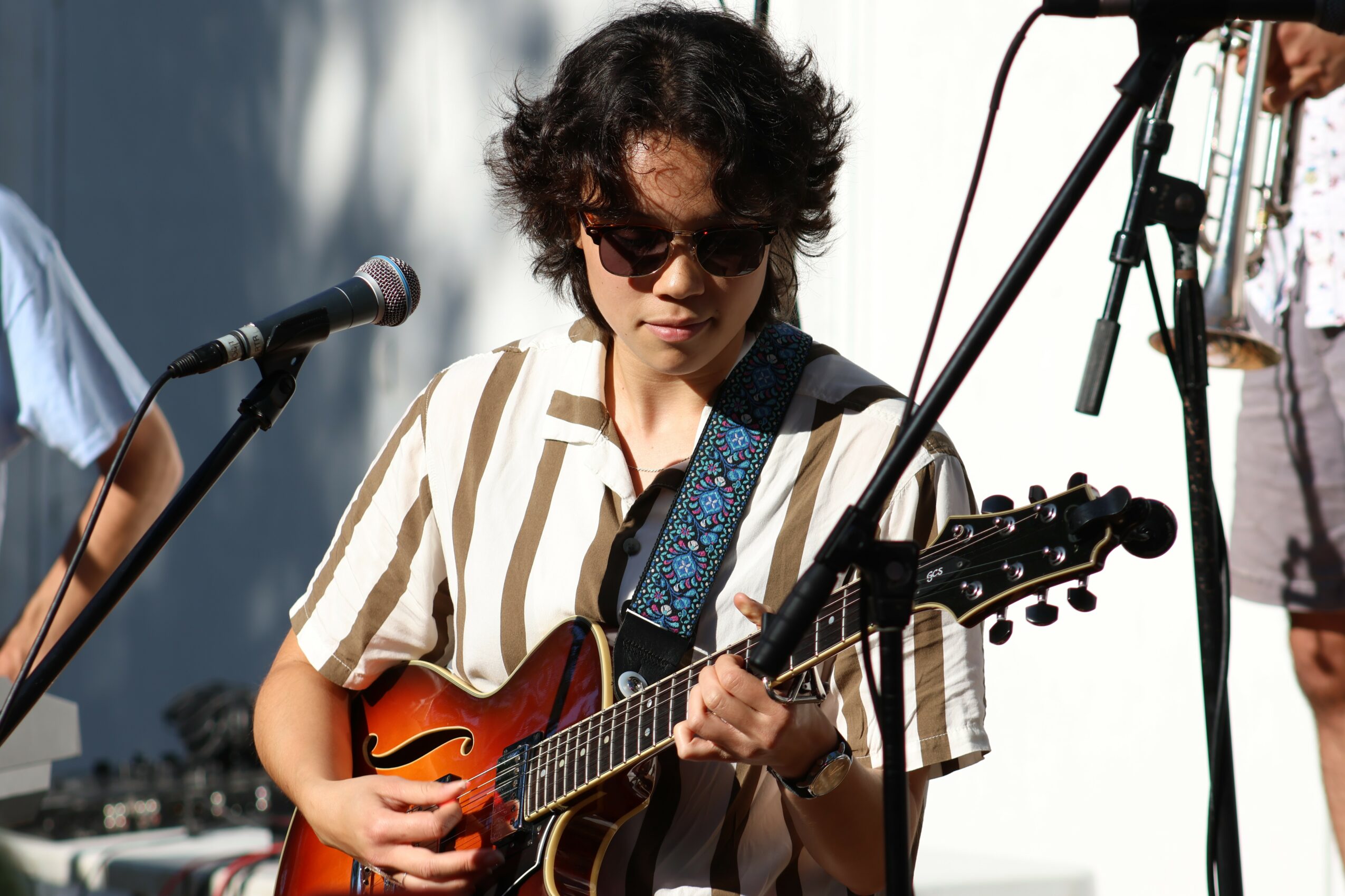 Person with curly hair wearing sunglasses and striped shirt playing guitar on stage with microphones.