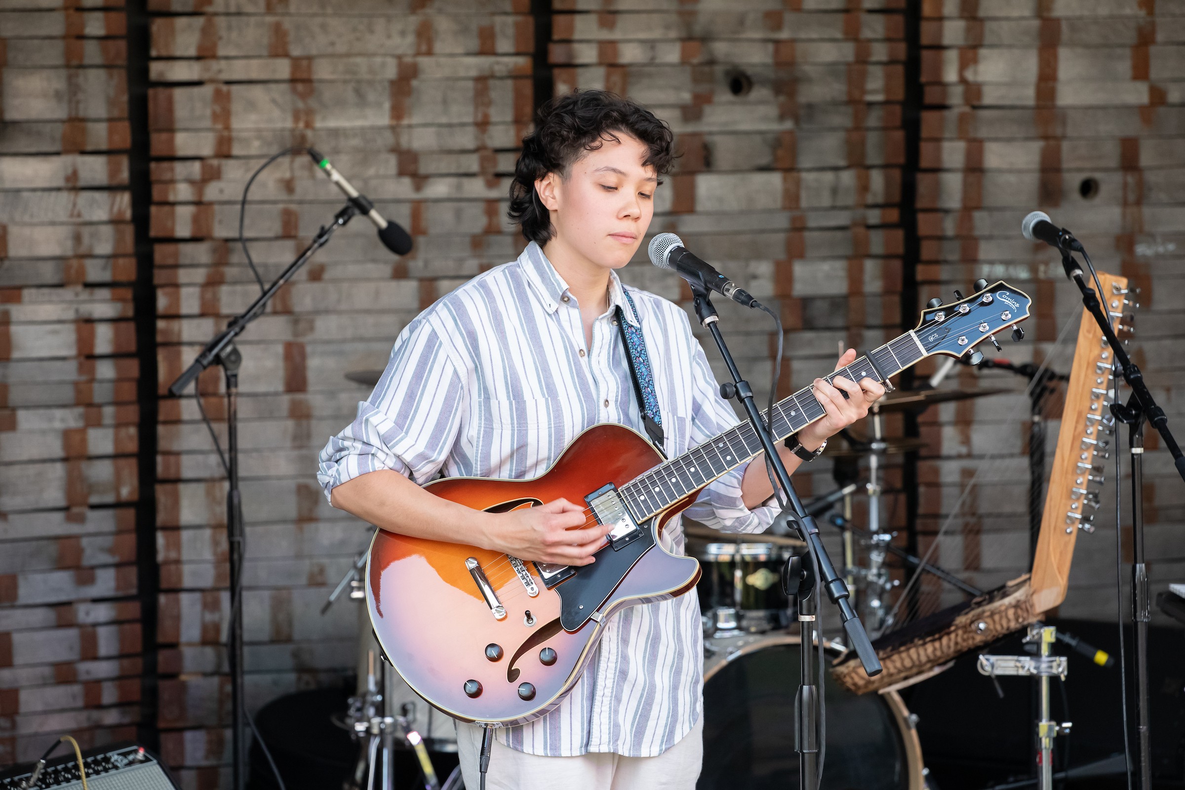Young person playing guitar and singing into a microphone on stage with brick wall background.