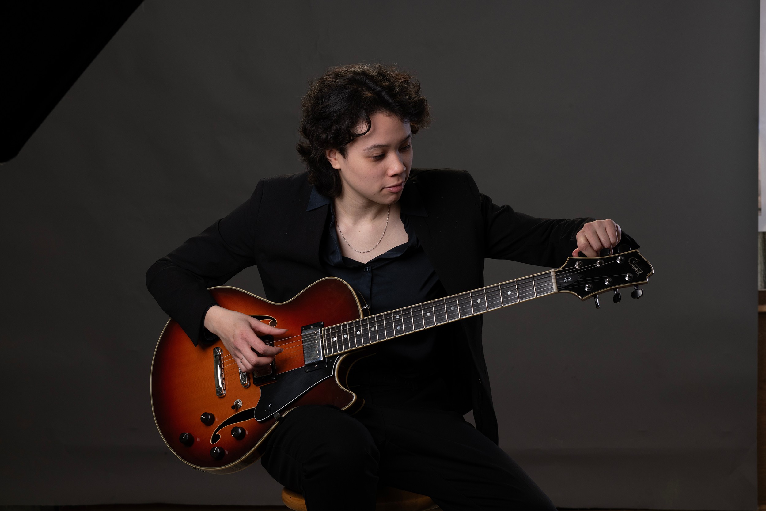 Person with short curly hair playing an electric guitar against a dark background.