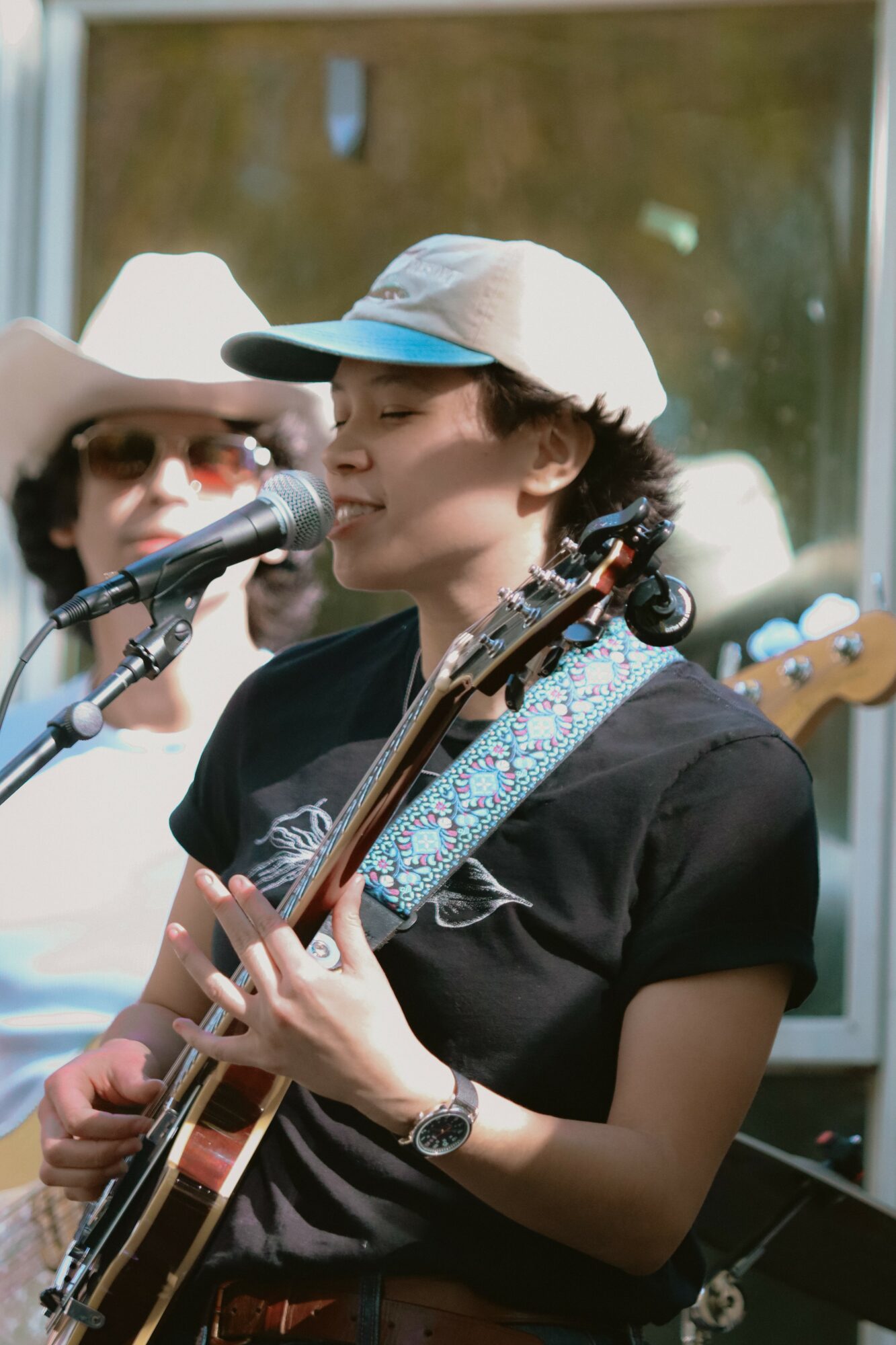 Young woman with short hair, wearing a white cap, playing guitar and singing into a microphone outdoors.