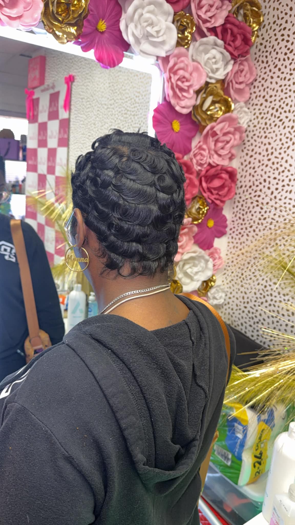 Back of a person with short, curly hair, wearing a black hoodie, in front of a floral display with pink, white, and gold flowers.