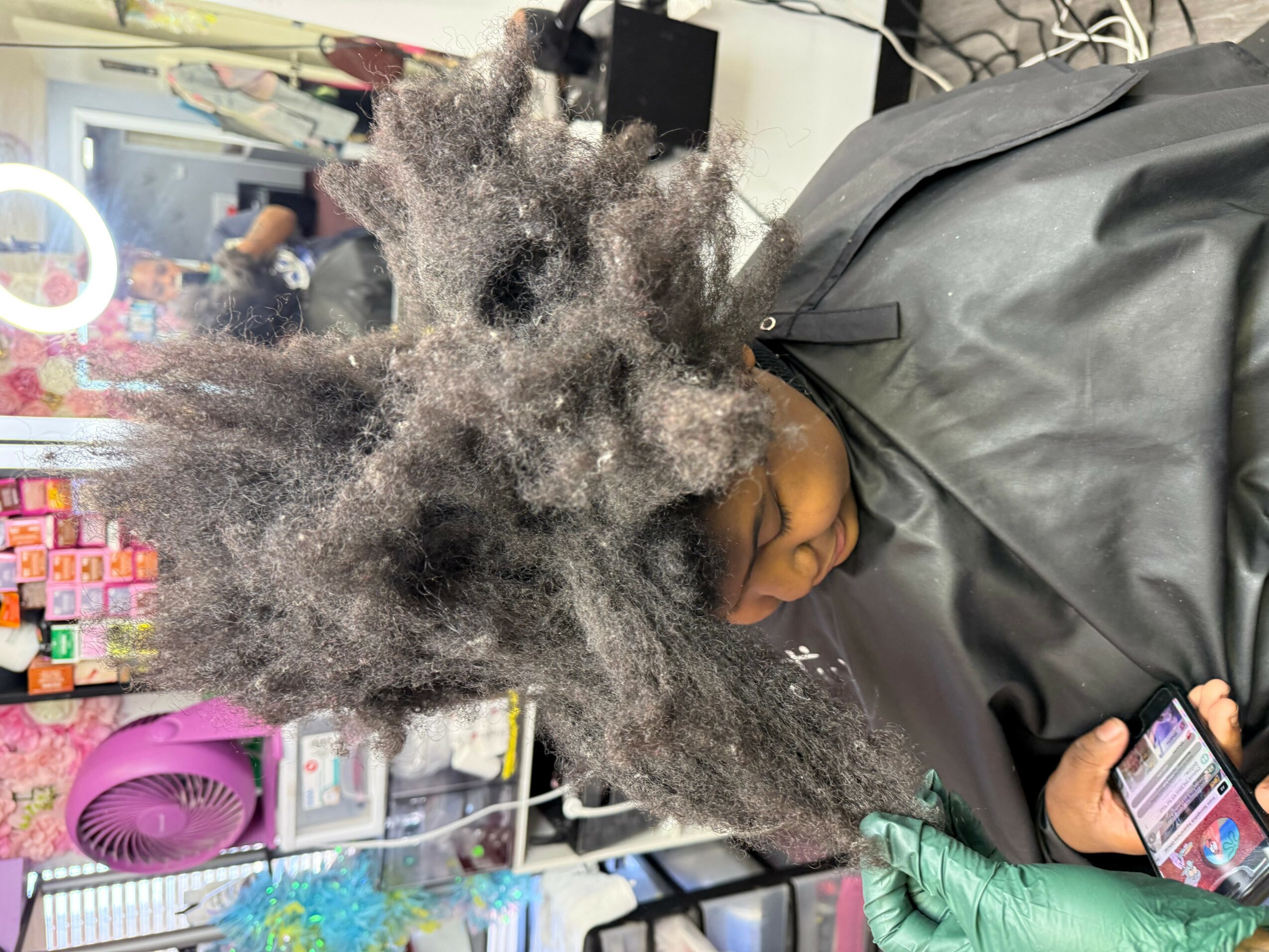Person with curly, frizzy hair sitting in a salon chair, looking at a phone, with salon products in the background.