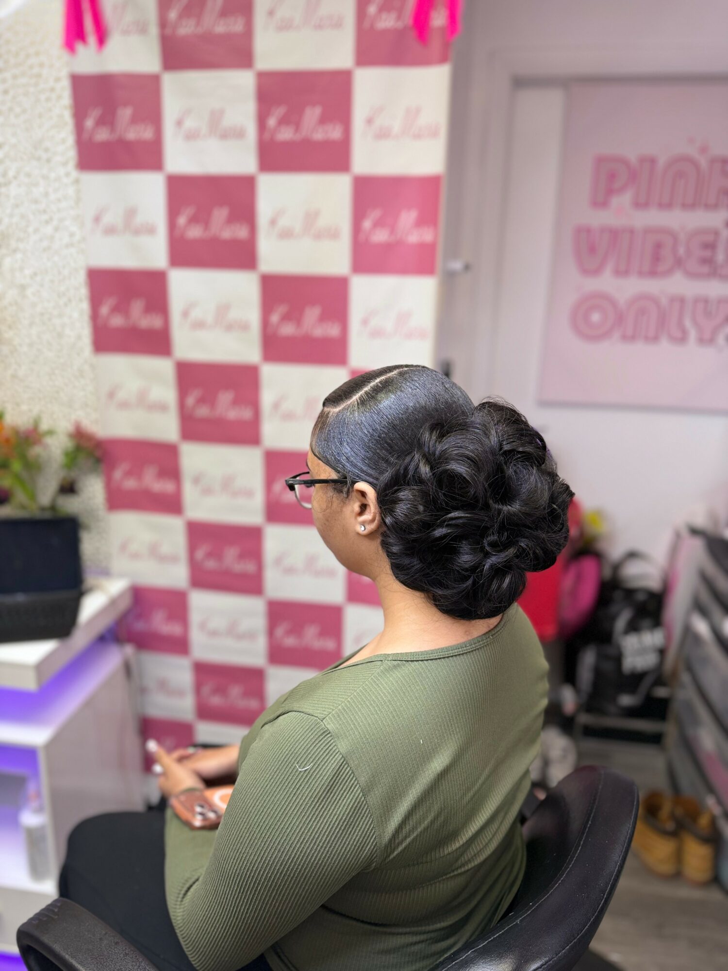 Woman with dark hair in a bun, wearing glasses and a green top, sitting indoors with pink and white checkered background.