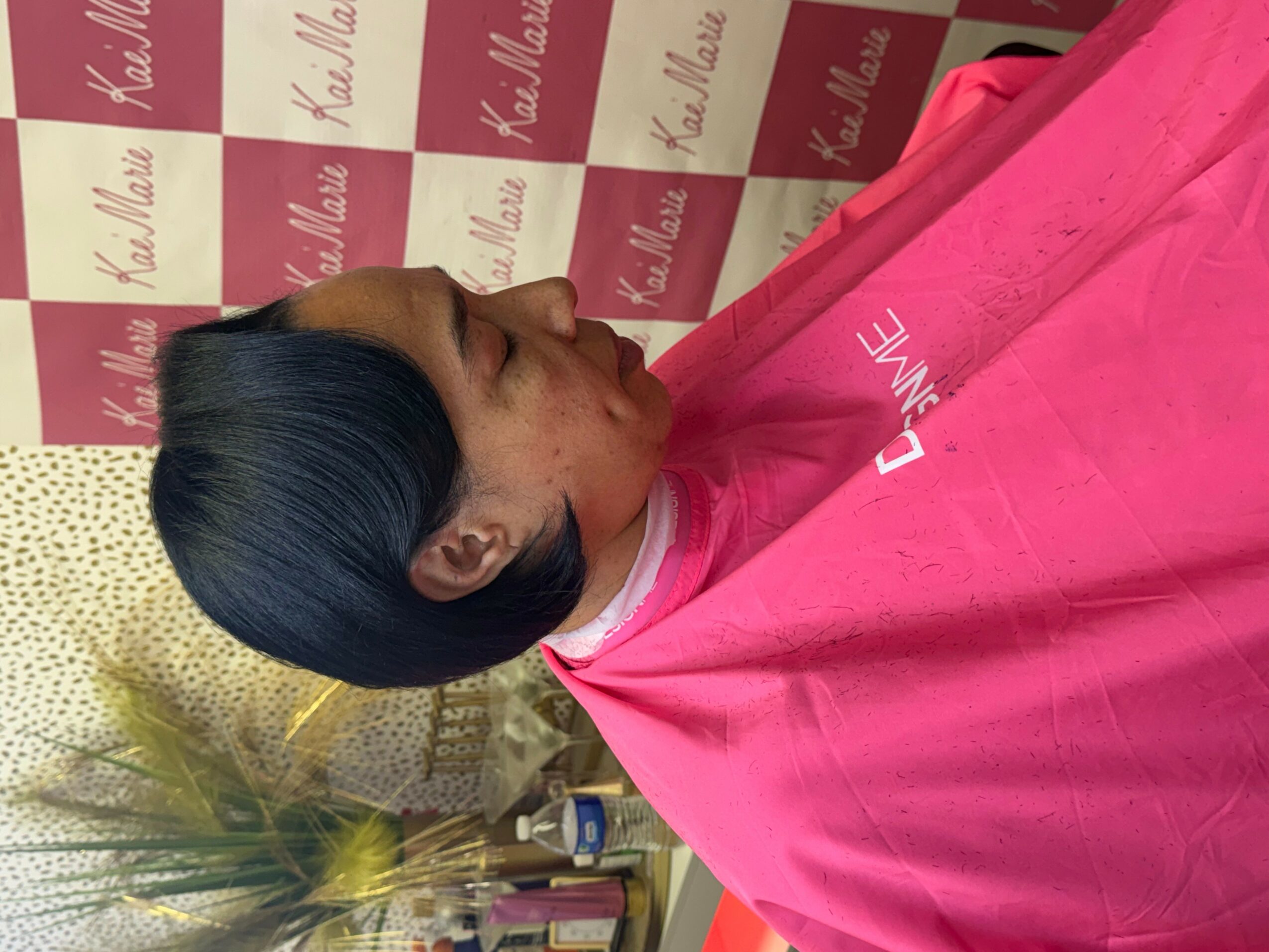 Person with short black hair in a pink cape, facing left, in a salon setting with pink and white background.