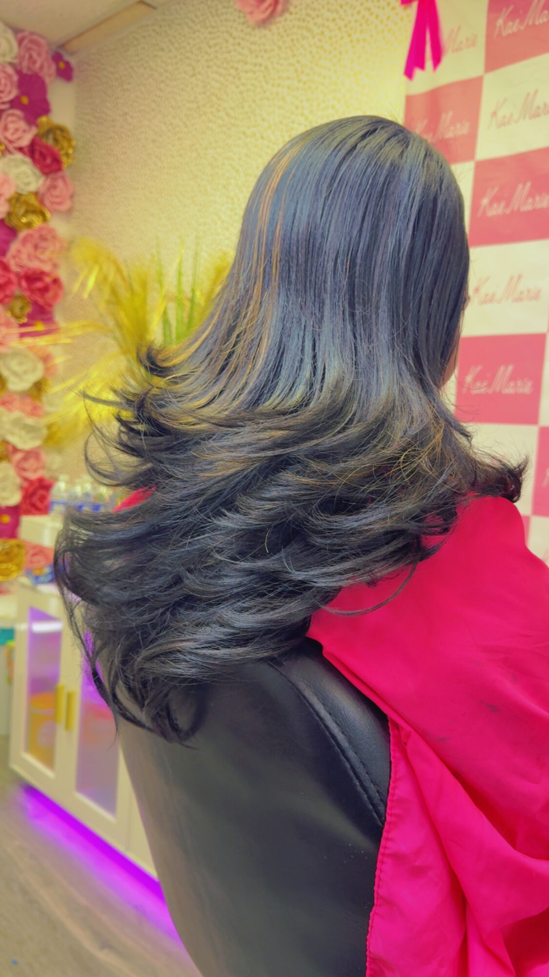 Back view of a woman with long, wavy black hair, wearing a red jacket, in front of a pink and white patterned background.