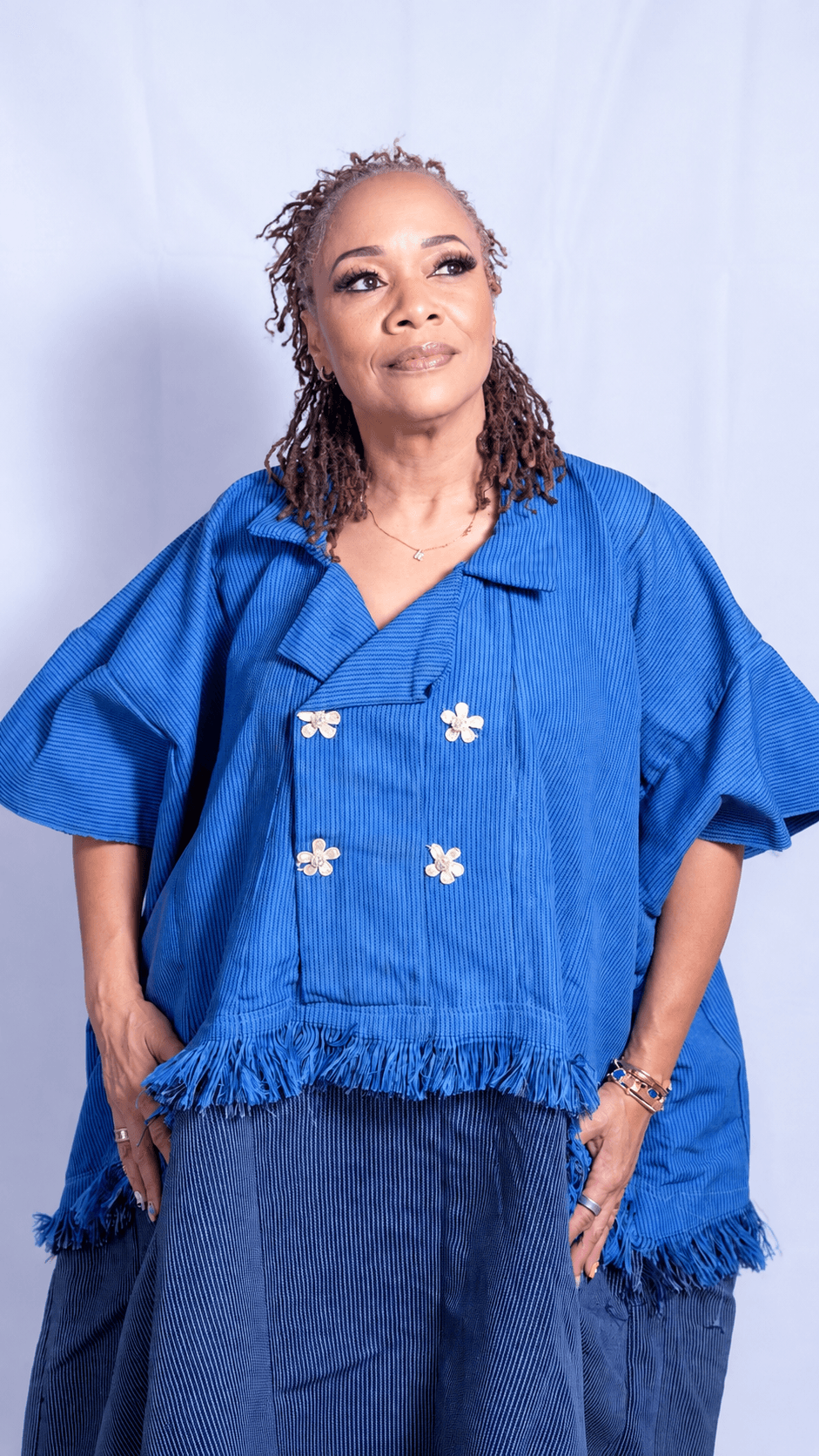 Woman with braided hair wearing a blue top with white buttons and denim pants, standing against a white background.