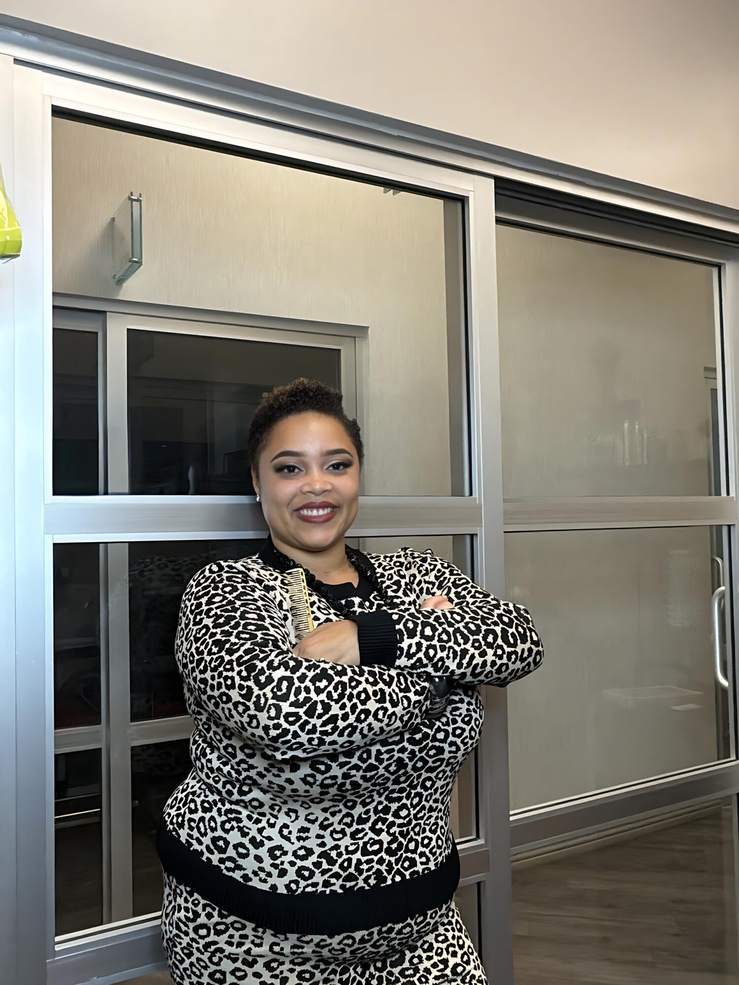 Smiling woman with crossed arms standing in front of glass windows, wearing a leopard print top and black pants.