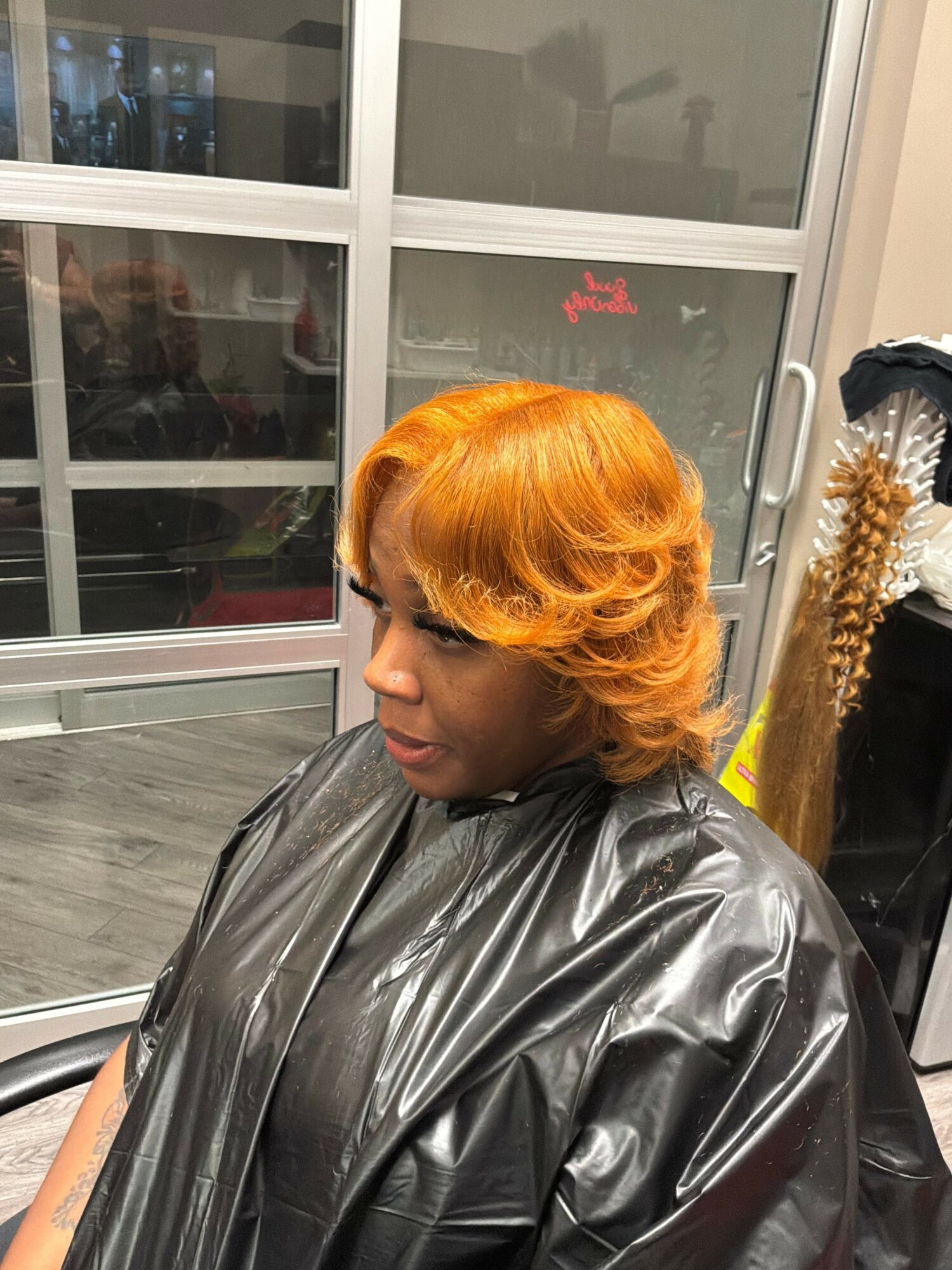 Woman with bright orange hair in a black plastic cape, looking down, in a salon or similar setting.