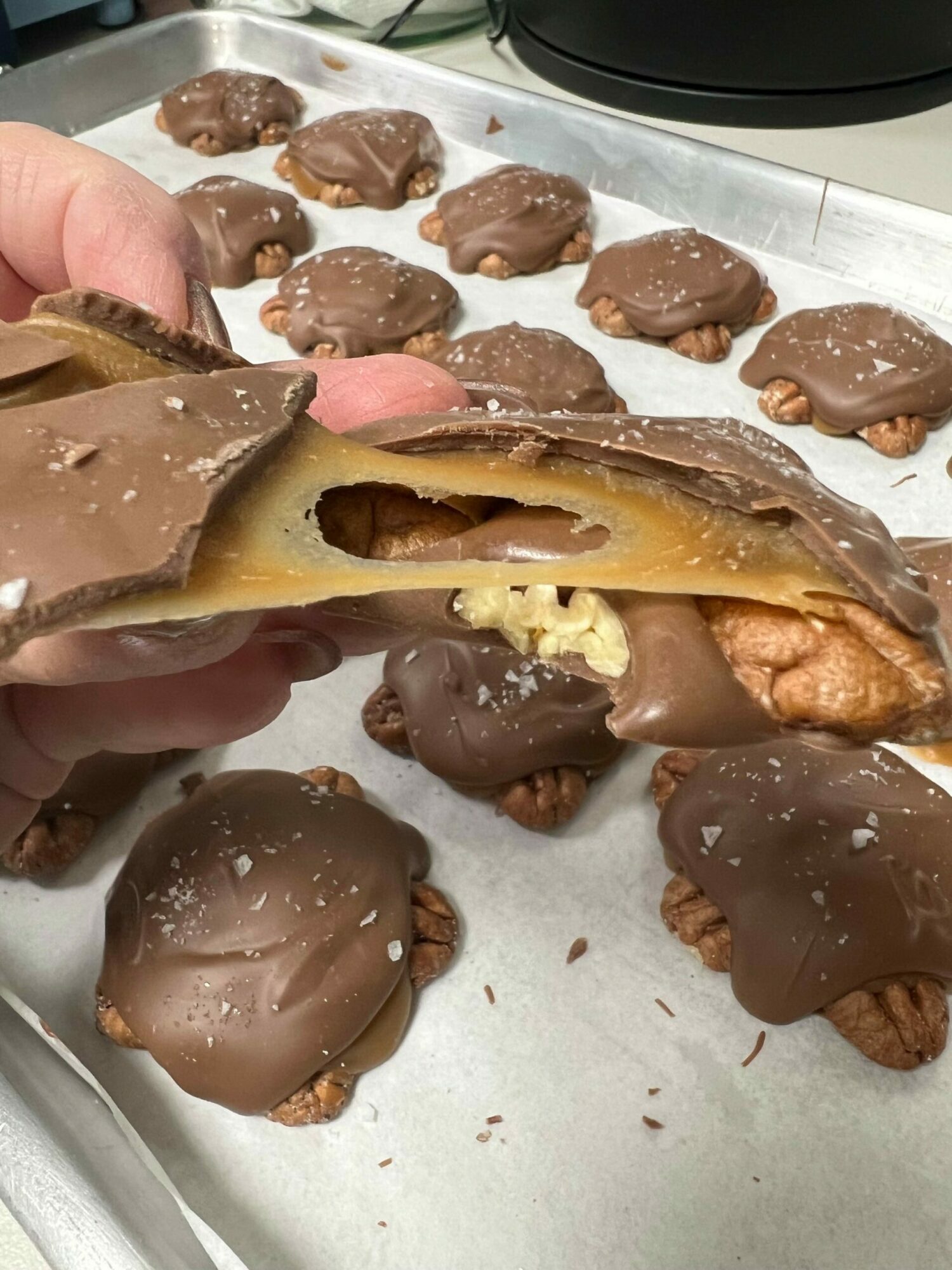 Hand holding a caramel-filled chocolate-covered treat with other similar confections on a tray, some with nuts or toppings, in a bakery or shop.