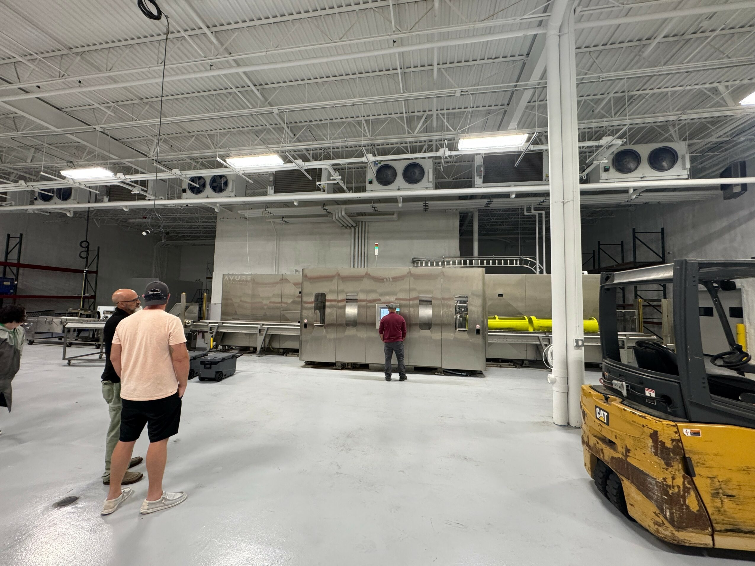Three people in a warehouse with equipment and a forklift, facing a large machine or storage unit, industrial setting.