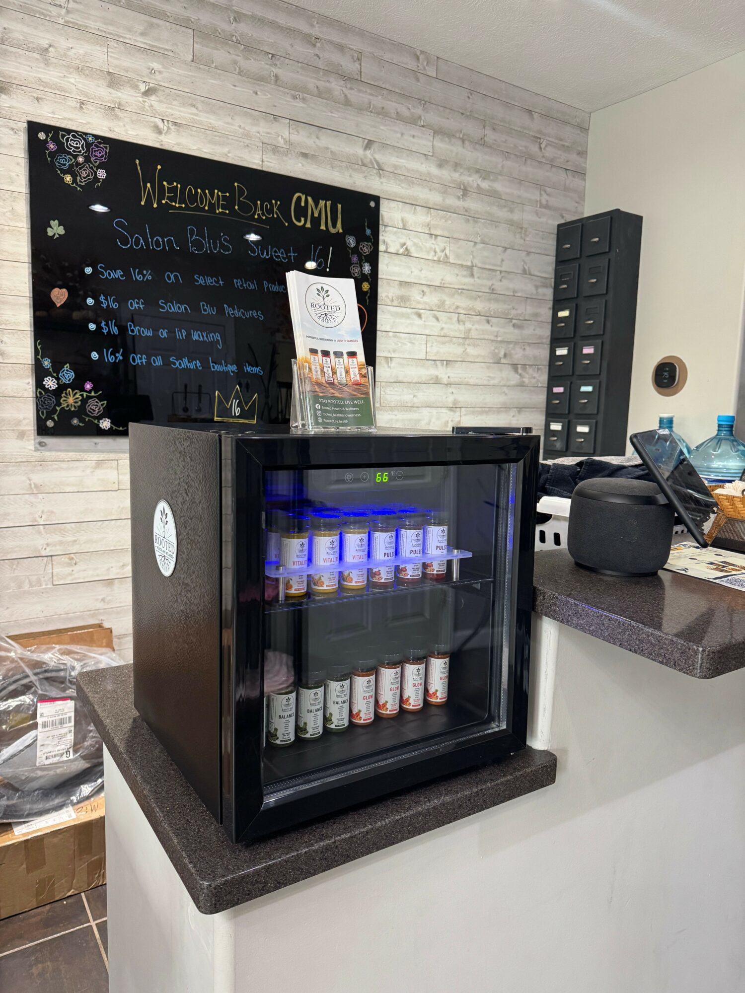 Black beverage cooler with drinks inside, on a countertop in a room with a blackboard and storage unit, viewed sideways.