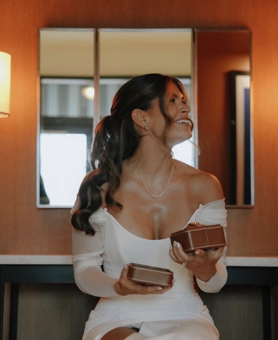 Woman in white dress smiling, holding a box, standing in front of a mirror with warm lighting.