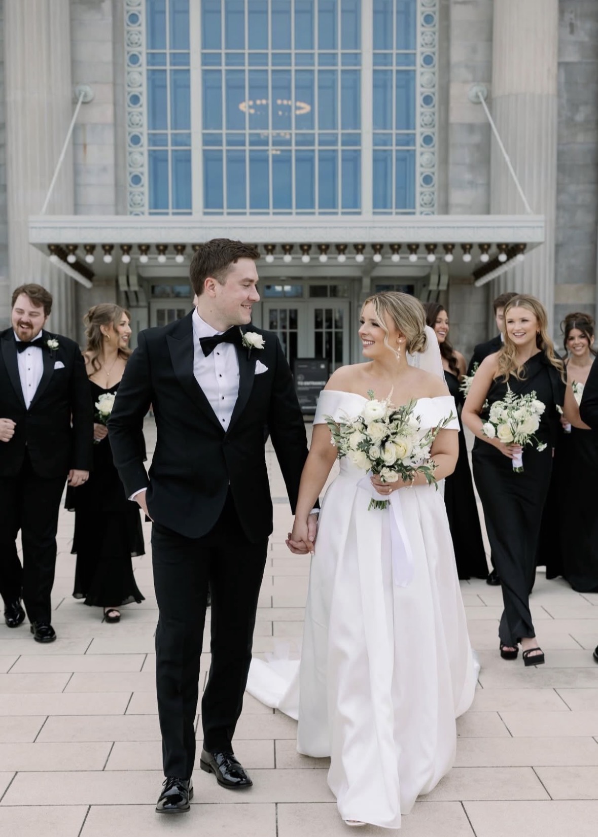 A wedding couple walking hand in hand outside a modern building, surrounded by bridesmaids and groomsmen in formal attire.