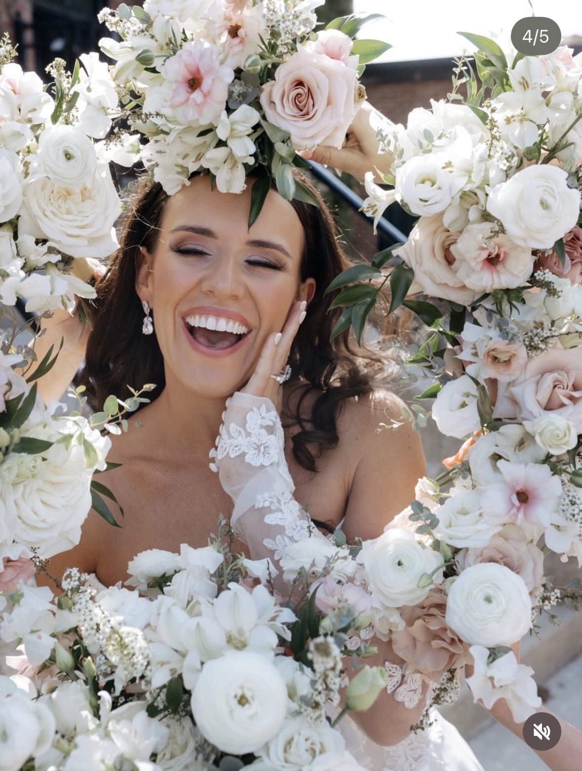 Woman with dark hair smiling surrounded by white and pink flowers, wearing earrings and a lace glove on her hand.
