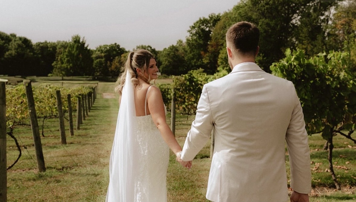A bride and groom holding hands outdoors in a vineyard, facing each other with trees and sky in the background.