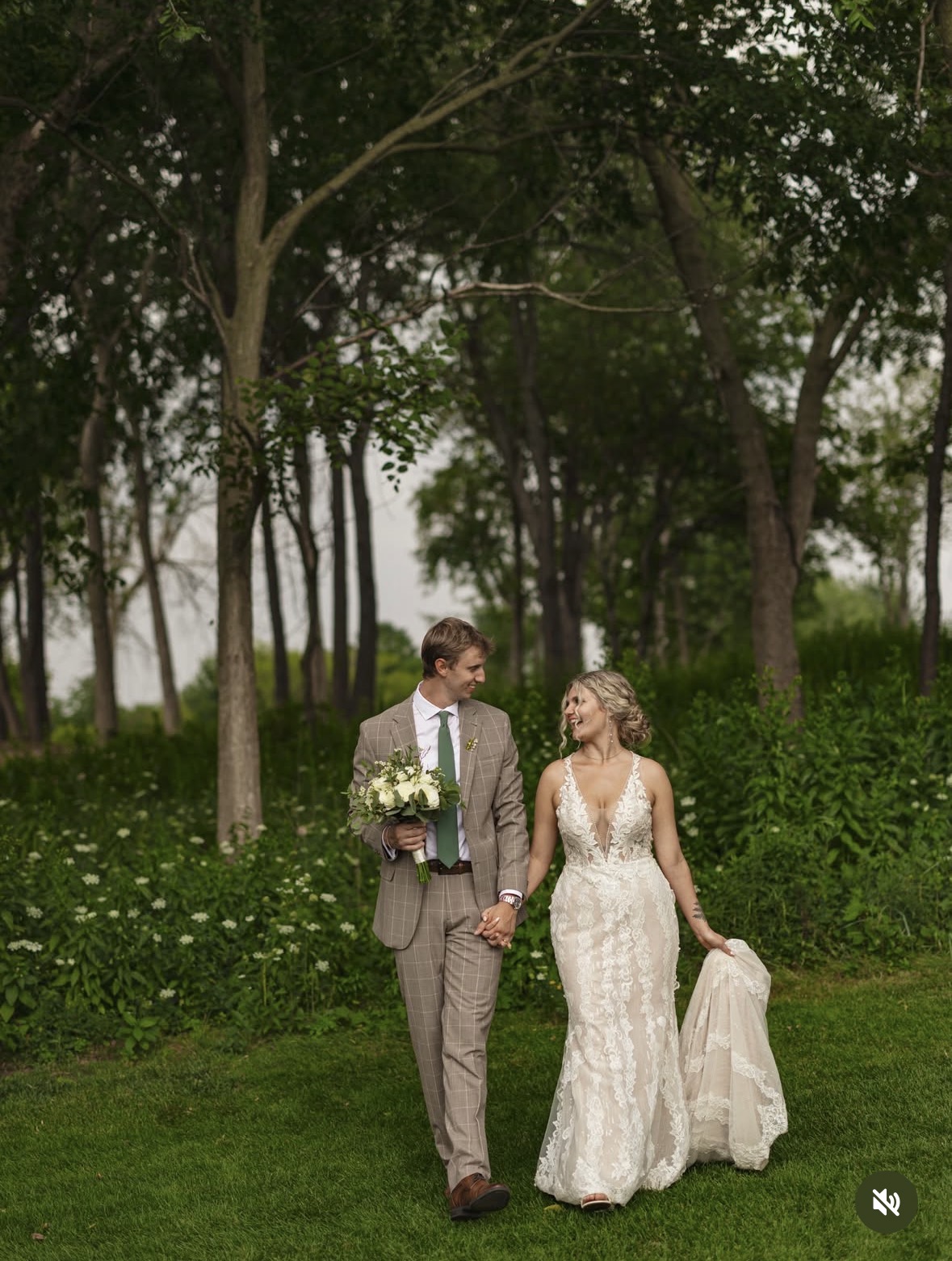 A couple holding hands outdoors in a park with trees and greenery, dressed in wedding attire, walking on grass.