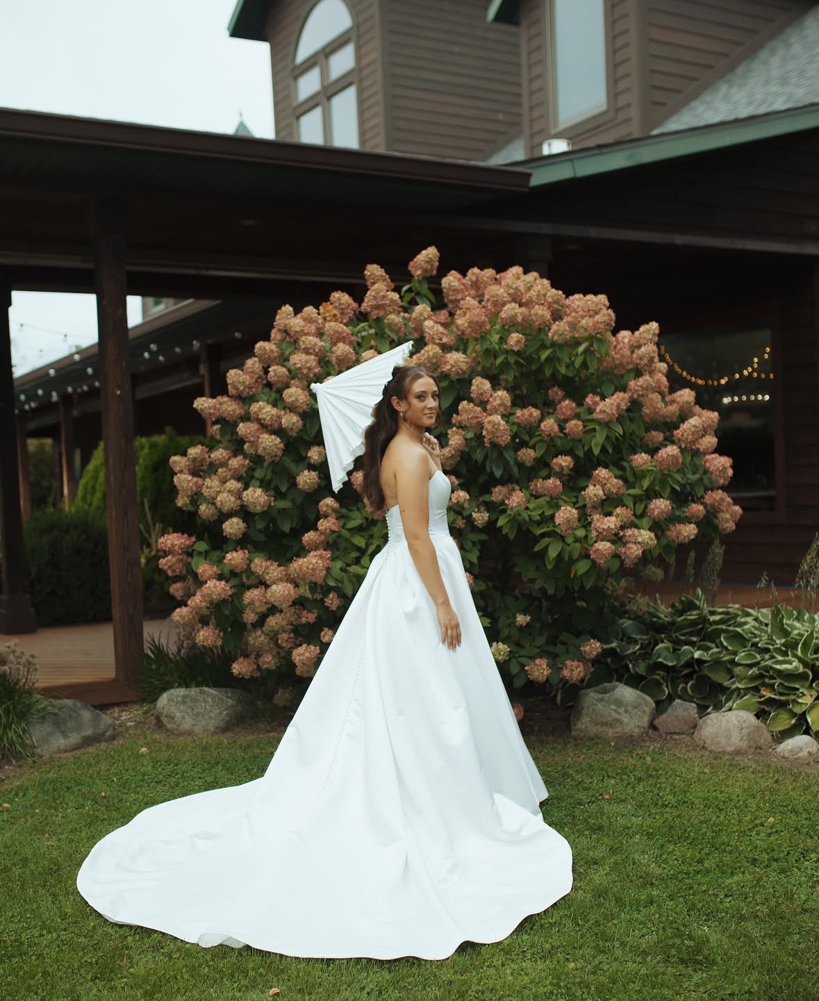 Woman in a white wedding dress standing outdoors near pink hydrangea bushes, holding a white parasol, with a house in the background.