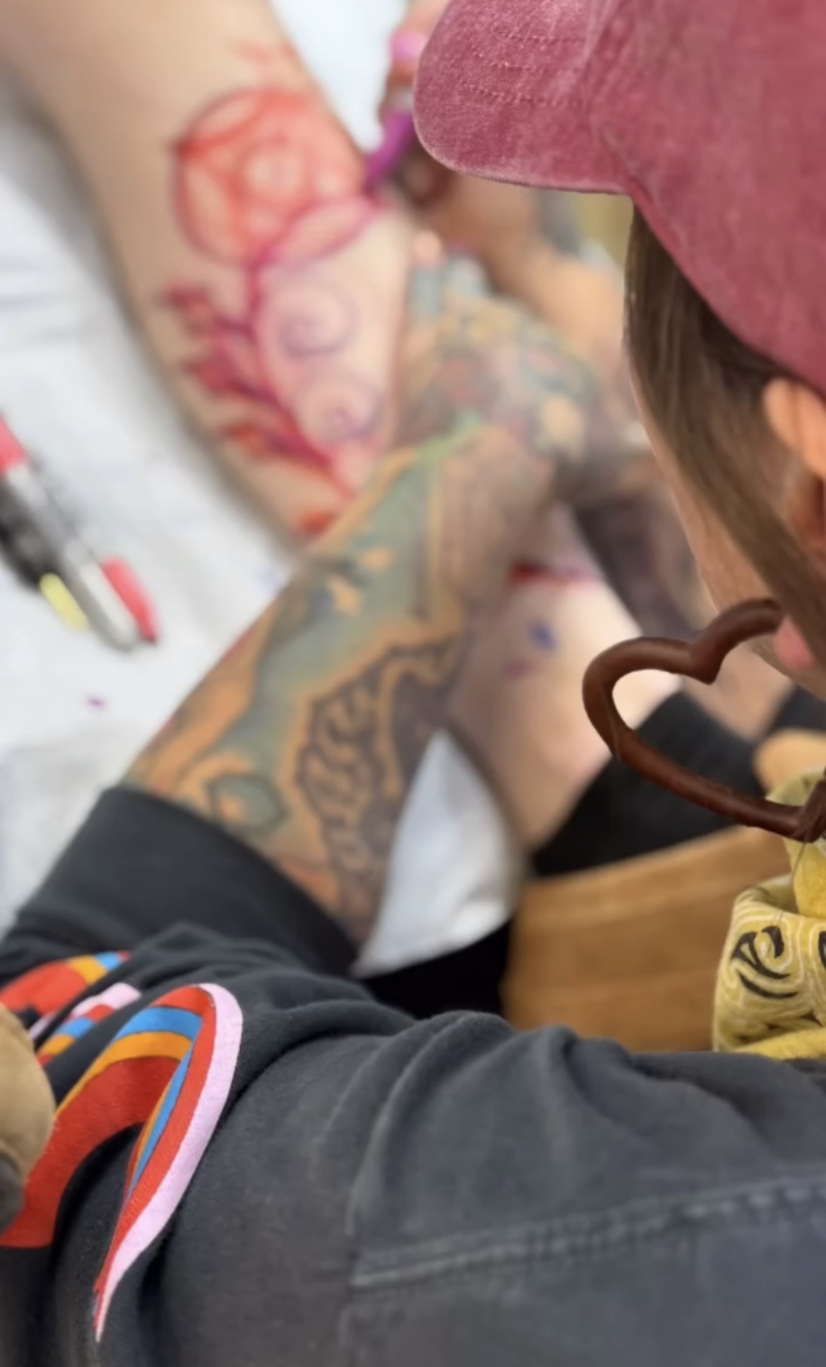 Person with tattoos on arm and hand, wearing glasses and a pink cap, in a tattooing process, with tattoo equipment nearby.