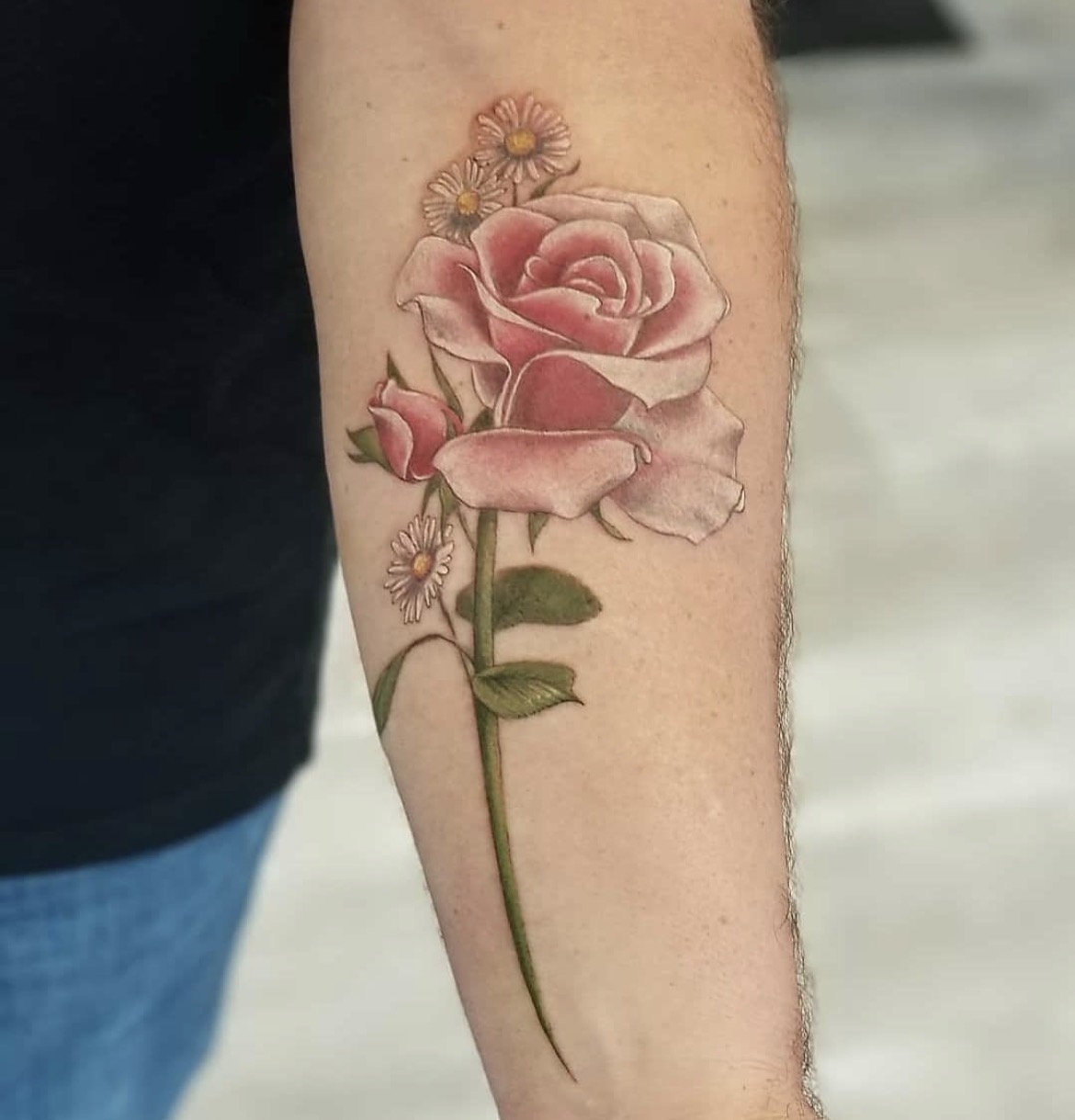 Tattoo of a pink rose with green leaves and small daisy-like flowers on a person's forearm.