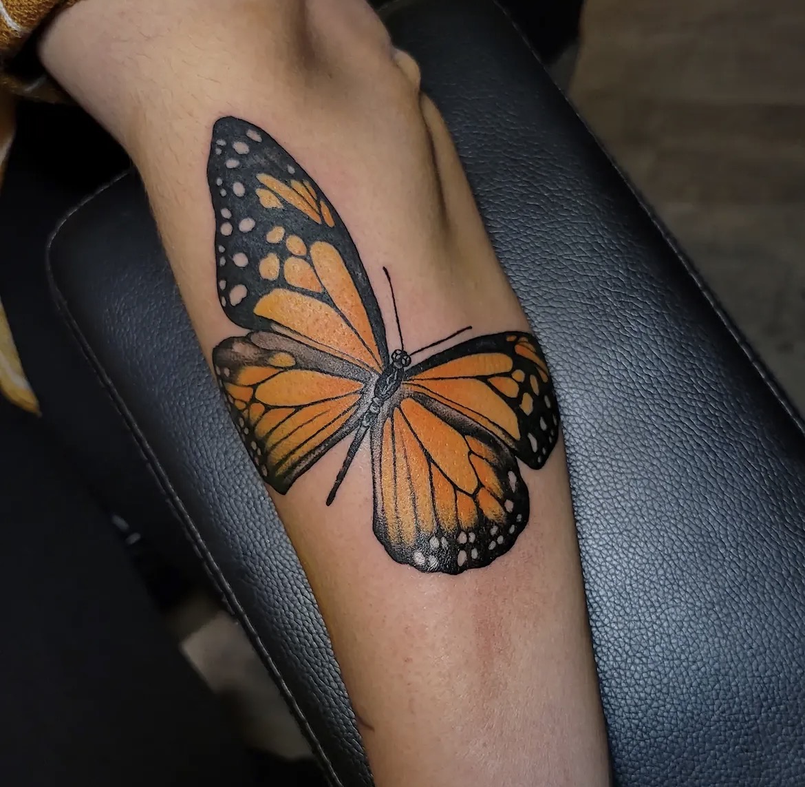 Colorful butterfly tattoo on person's arm, with orange, black, and white wings, resting on a black surface.