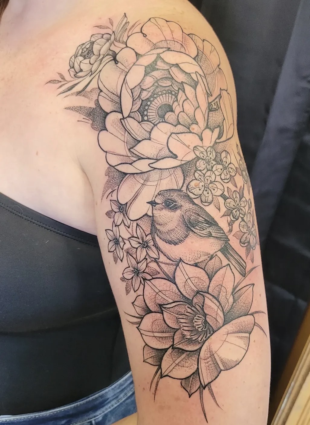 Tattoo of flowers and a bird on person's upper arm, detailed line work, black and gray shading.