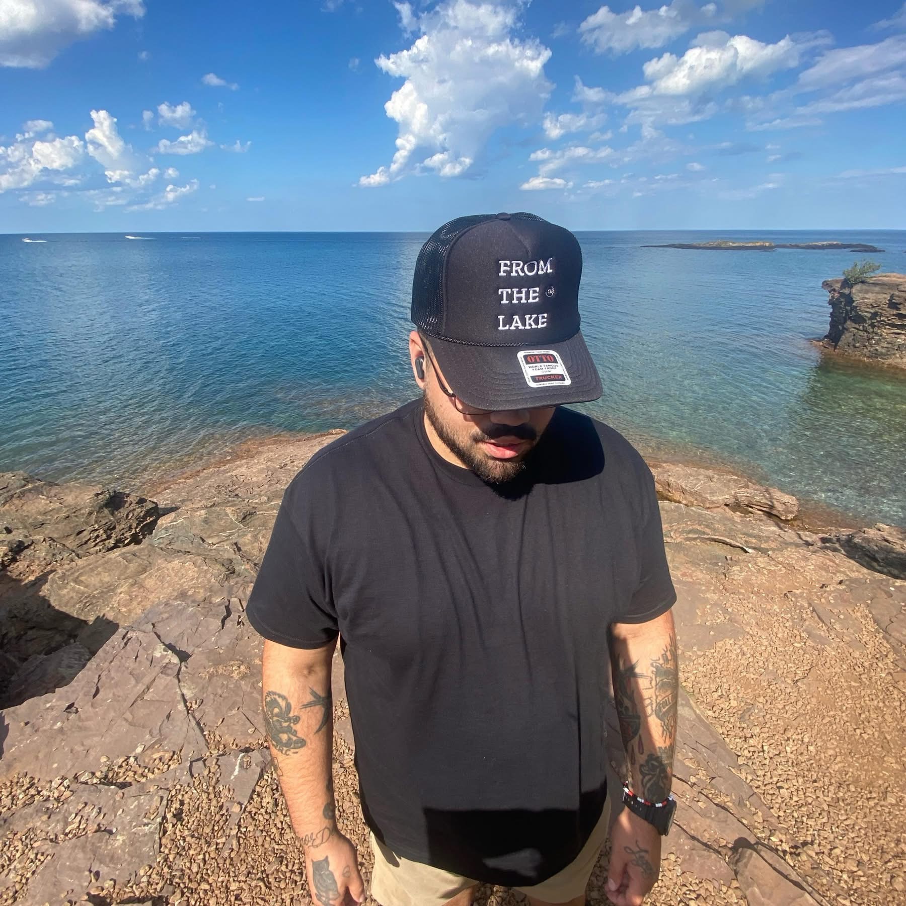 Man wearing a black cap and t-shirt stands on rocks near a lake with blue sky and clouds.