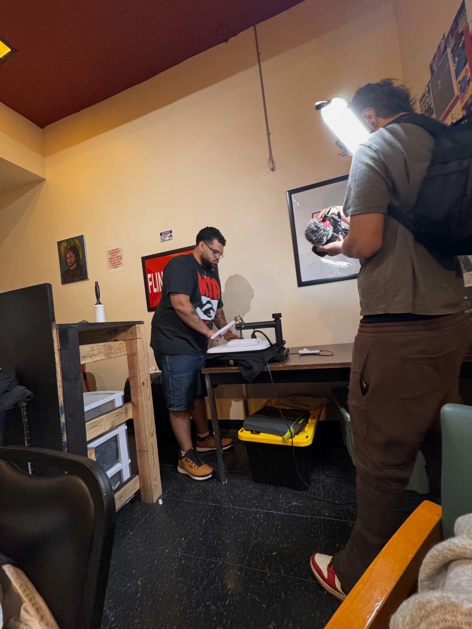 Two men in a room with artwork on the walls, one using a machine on a table, the other standing nearby with a backpack.