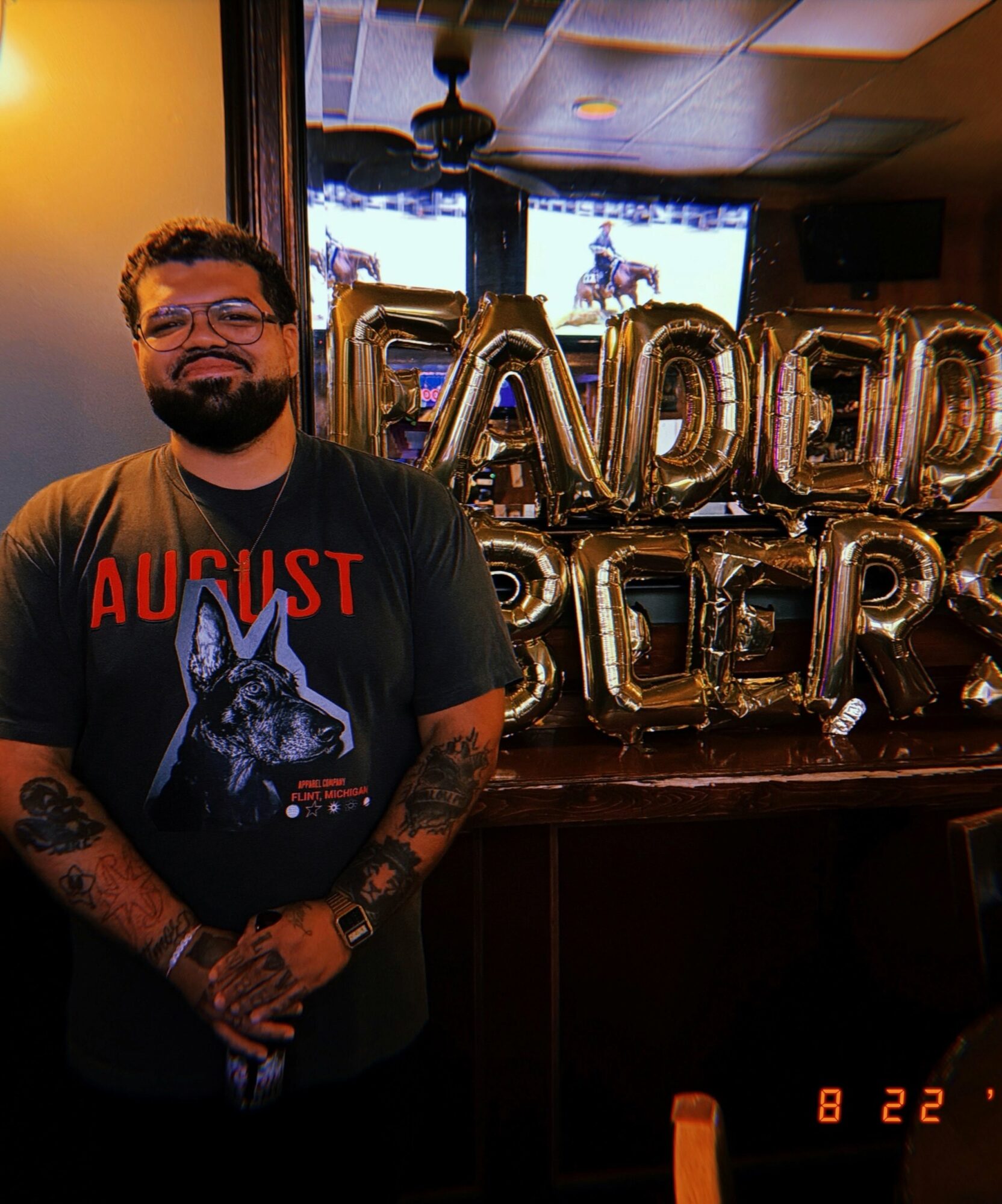 Man with glasses and beard smiling, wearing a black T-shirt with a wolf graphic, standing indoors with balloons in the background.
