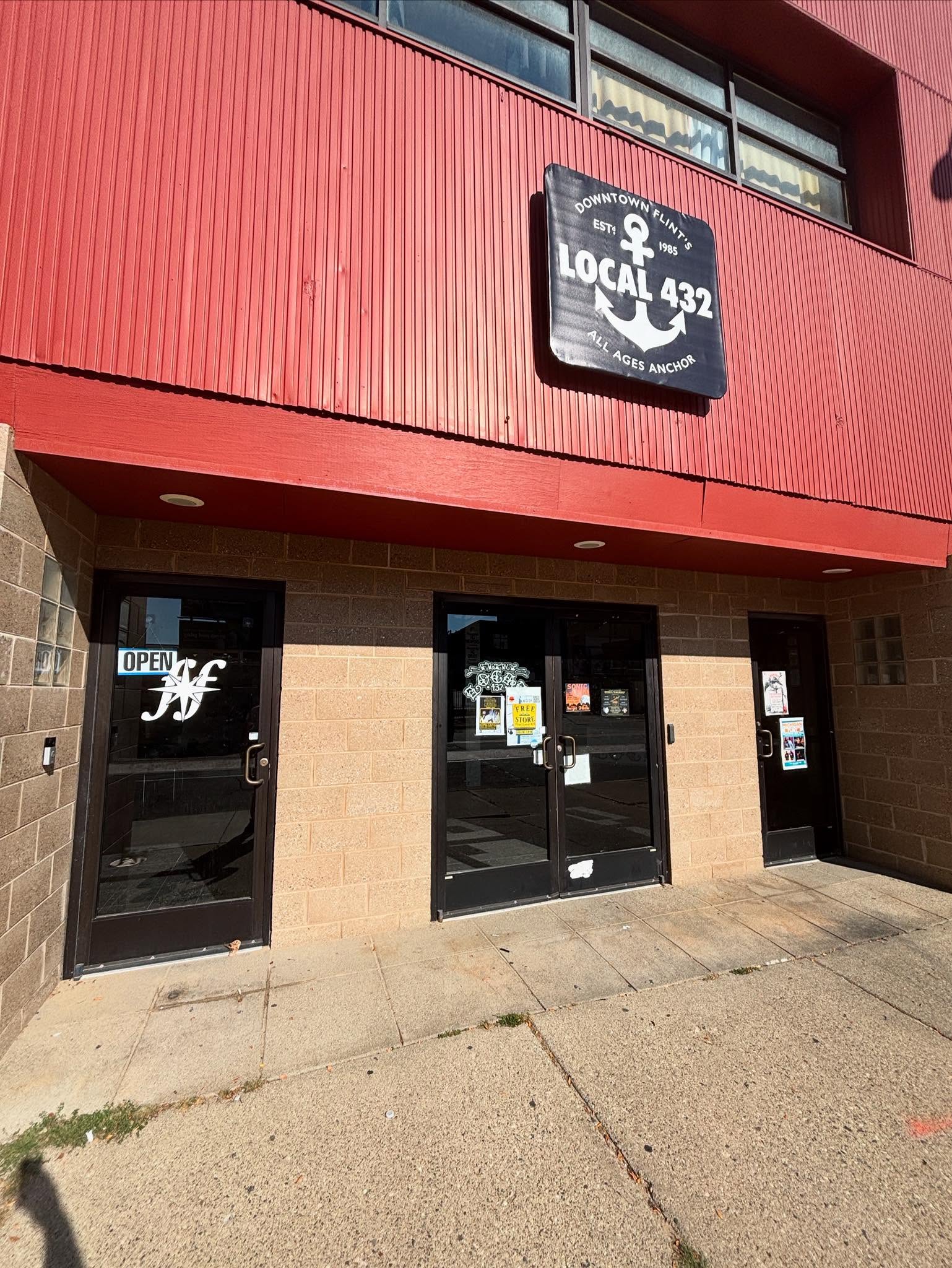 Building entrance with three glass doors, signs indicating open hours, and a sign for Local 432 union on the red upper wall.