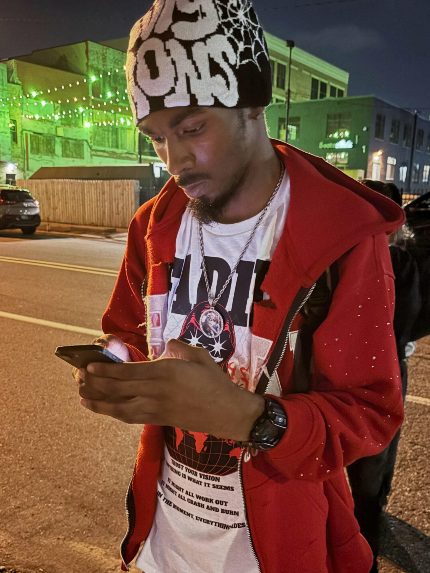 Young man wearing a beanie and red jacket looking at his phone on street at night.