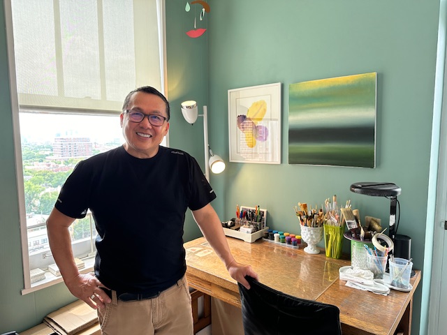 Smiling man standing in a room with a desk, colorful artwork, and a window showing a city view.
