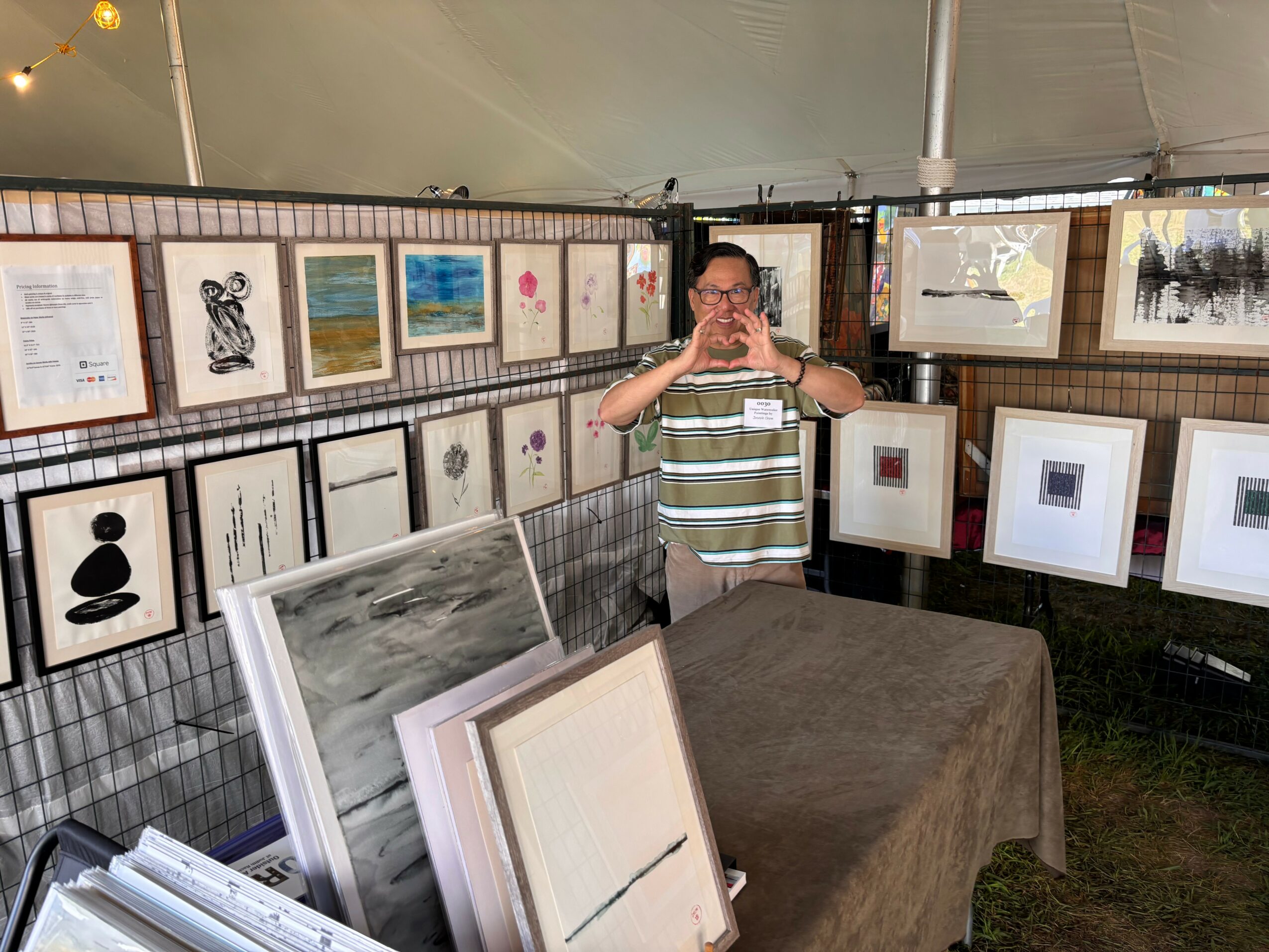 Person taking a photo in art display area, with framed artwork on walls and table with art pieces, outdoors.
