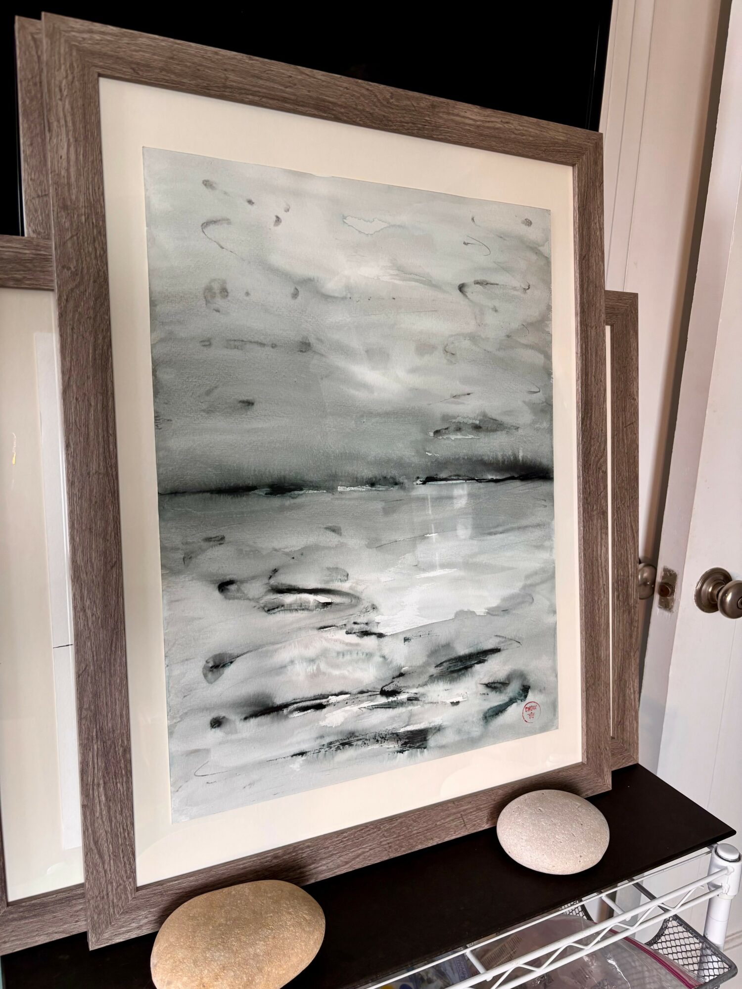 Abstract black, gray, and white watercolor painting in a wooden frame, leaning against a wall on a table with two stones.