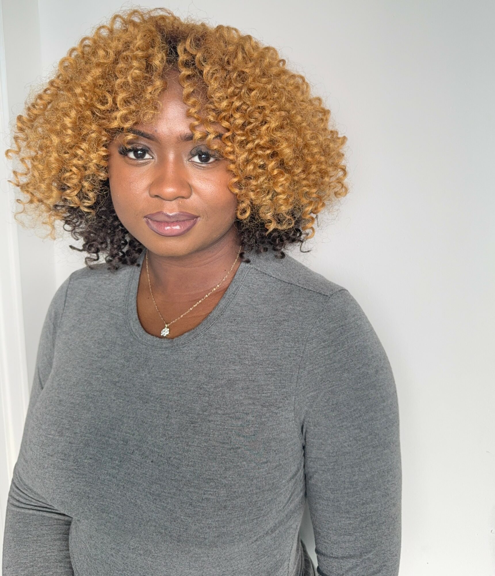 Young woman with curly blonde hair wearing a gray top, looking at the camera against a plain background.