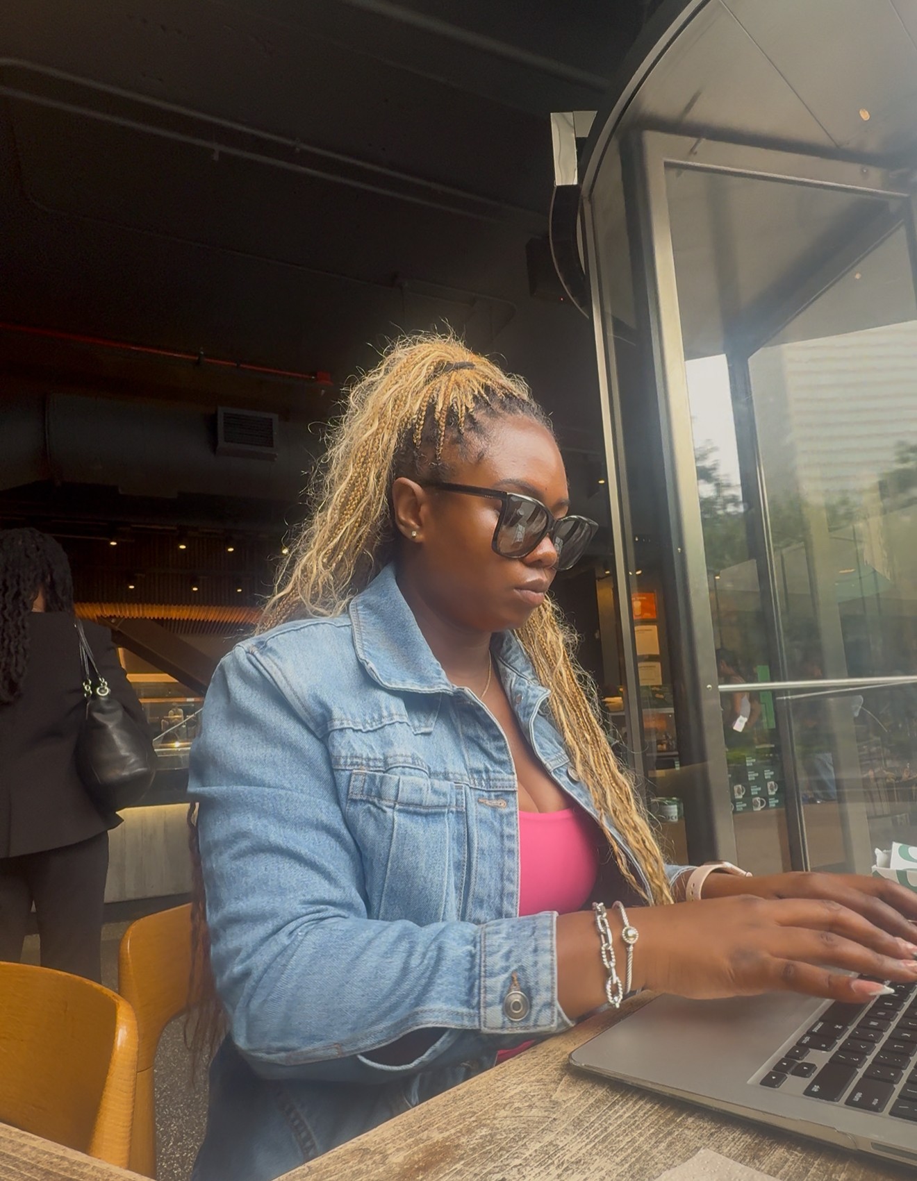 Woman with long curly hair, sunglasses, denim jacket, and pink top using a laptop at a table.