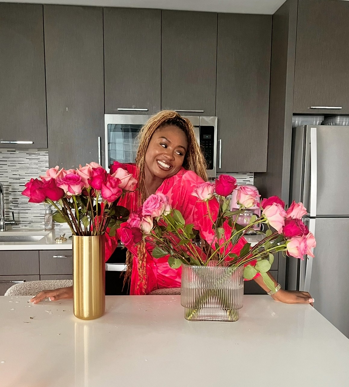 Woman smiling behind two vases of pink roses on kitchen counter.
